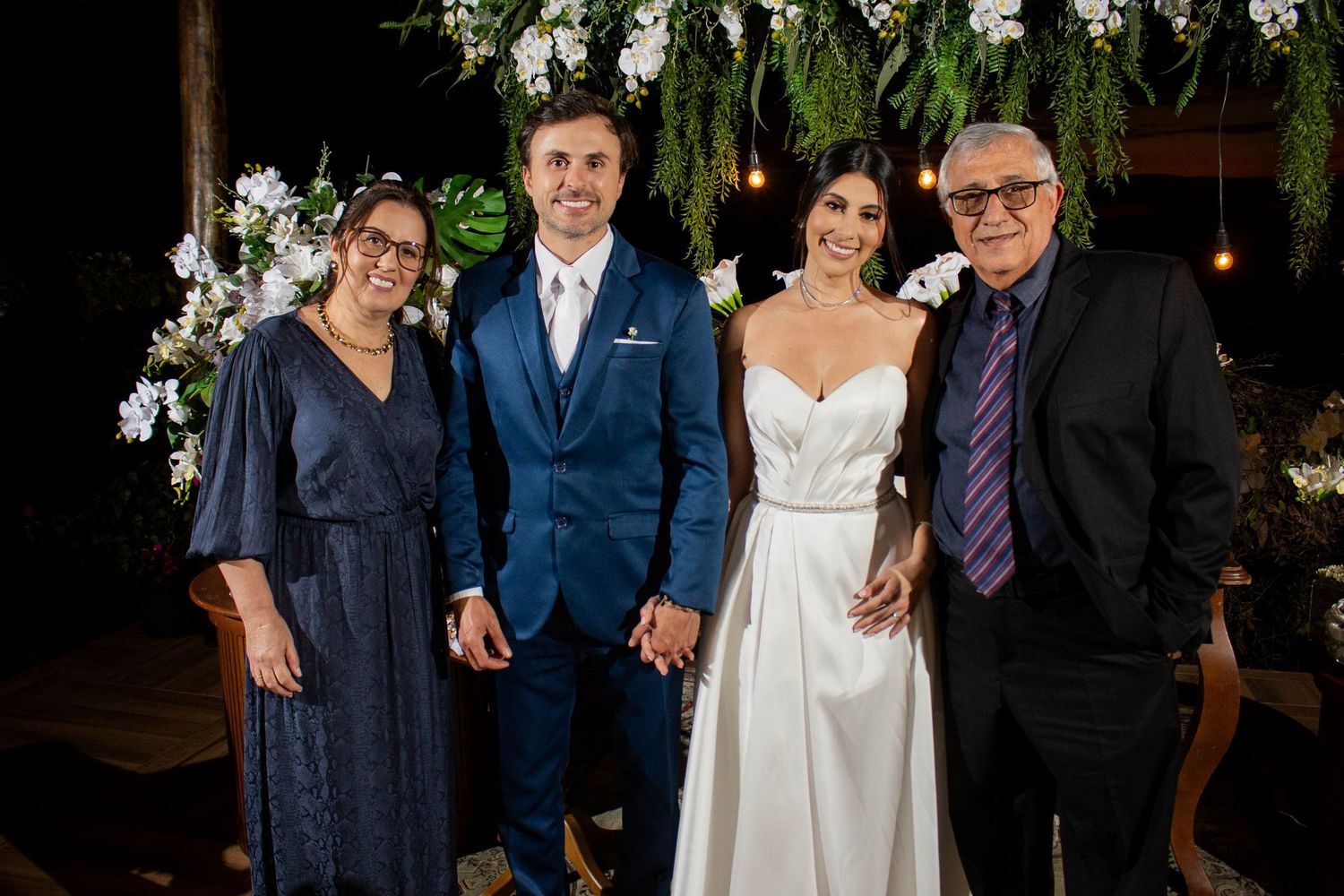 Wedding couple poses with family members in front of floral arch at elegant outdoor evening ceremony.