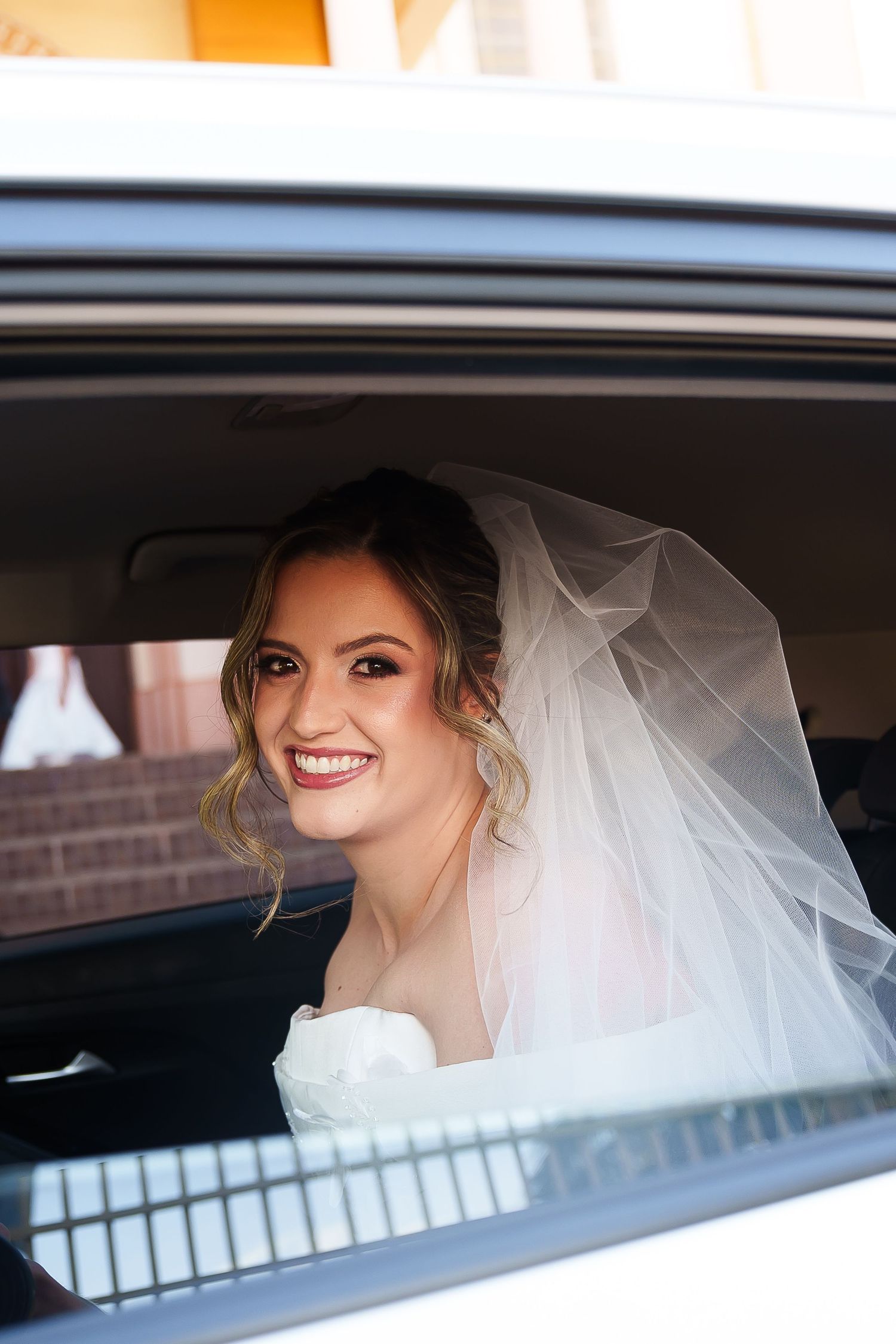 Radiant bride wearing veil smiling from car window on wedding day.
