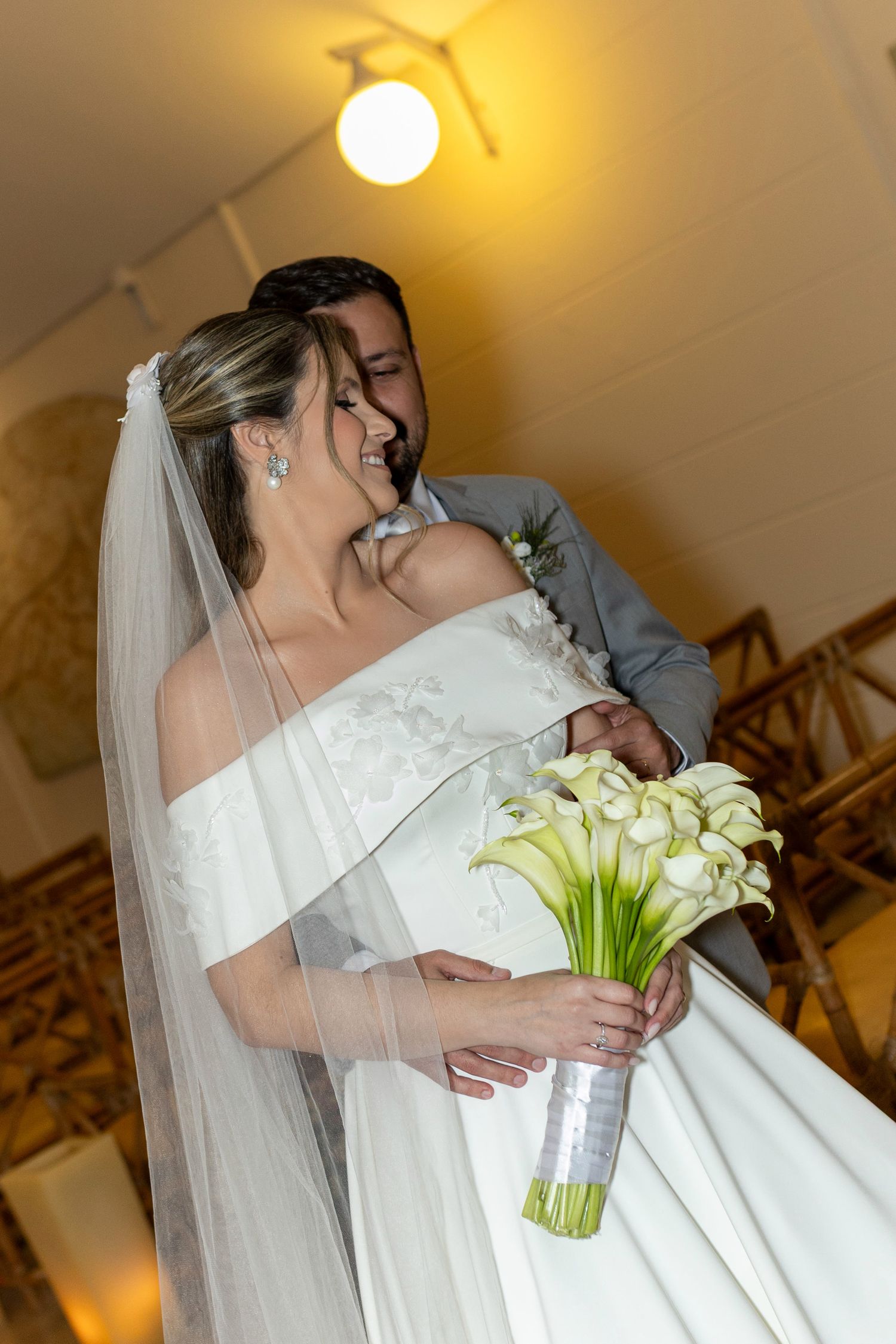 Bride in white gown holding calla lily bouquet embraced by groom in gray suit, smiling joyfully indoors.