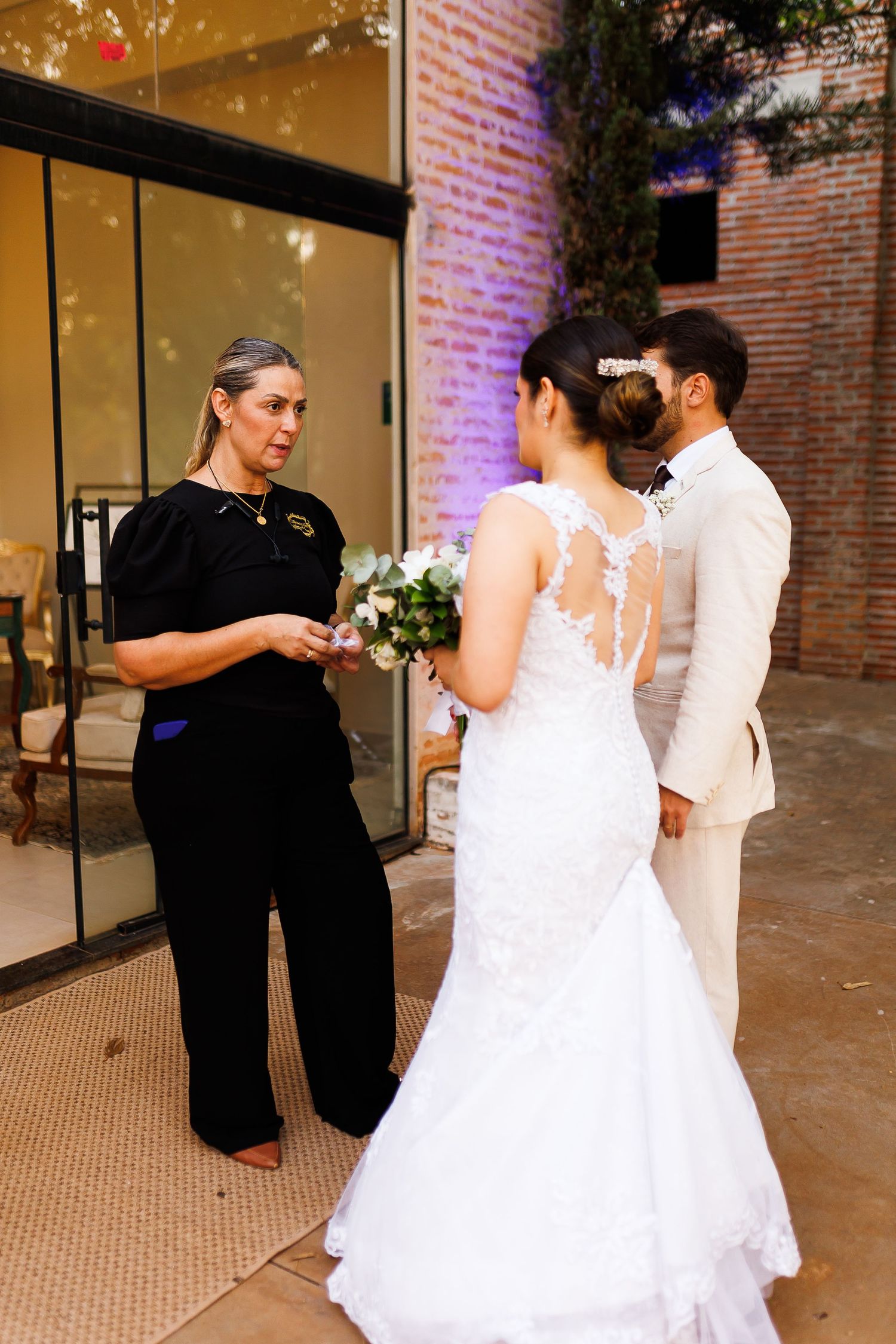 Bride in lace mermaid gown and groom in tan suit stand before officiant during outdoor wedding ceremony.