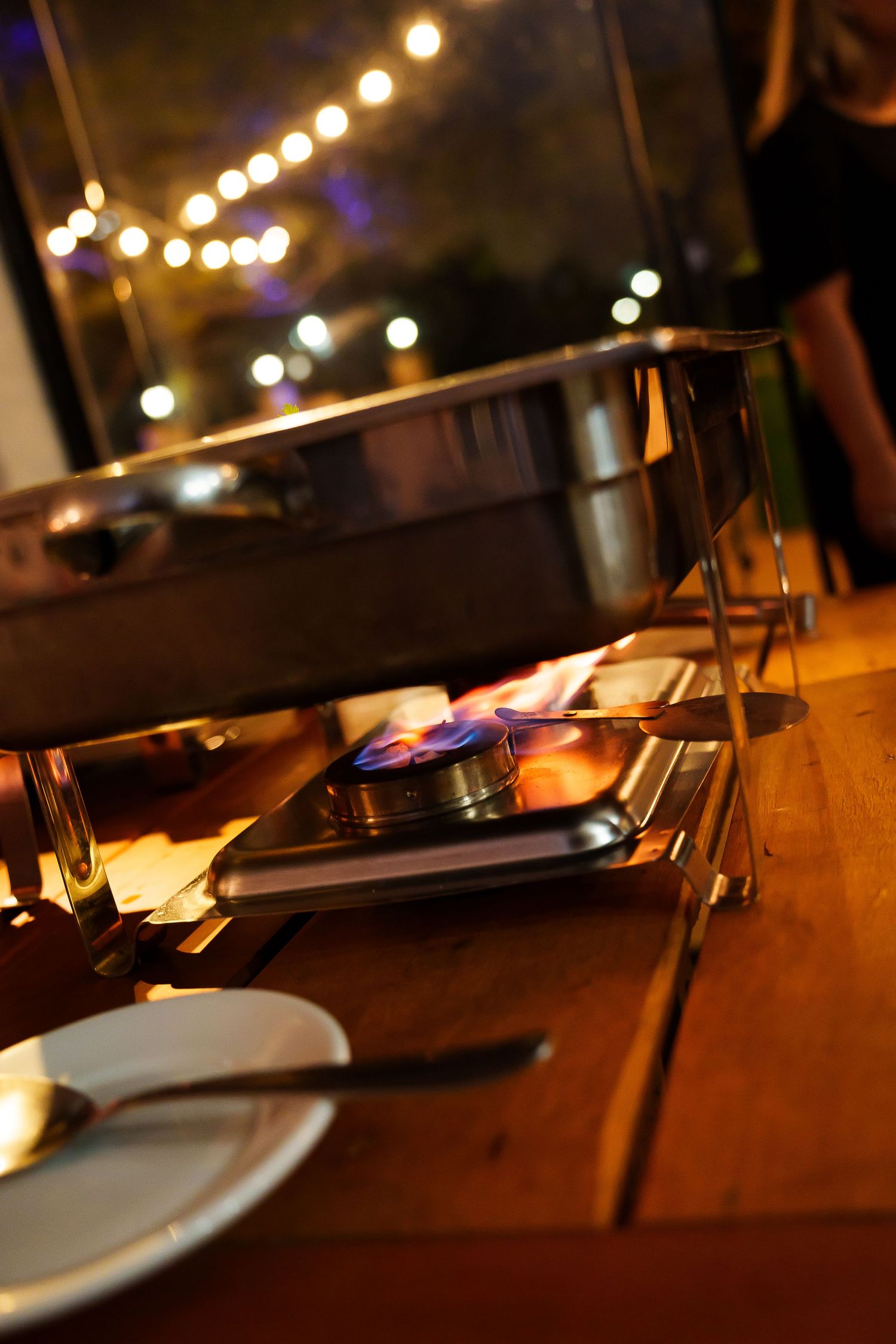 Chafing dishes with blue flames on a wooden buffet table at an outdoor evening event with string lights in background.