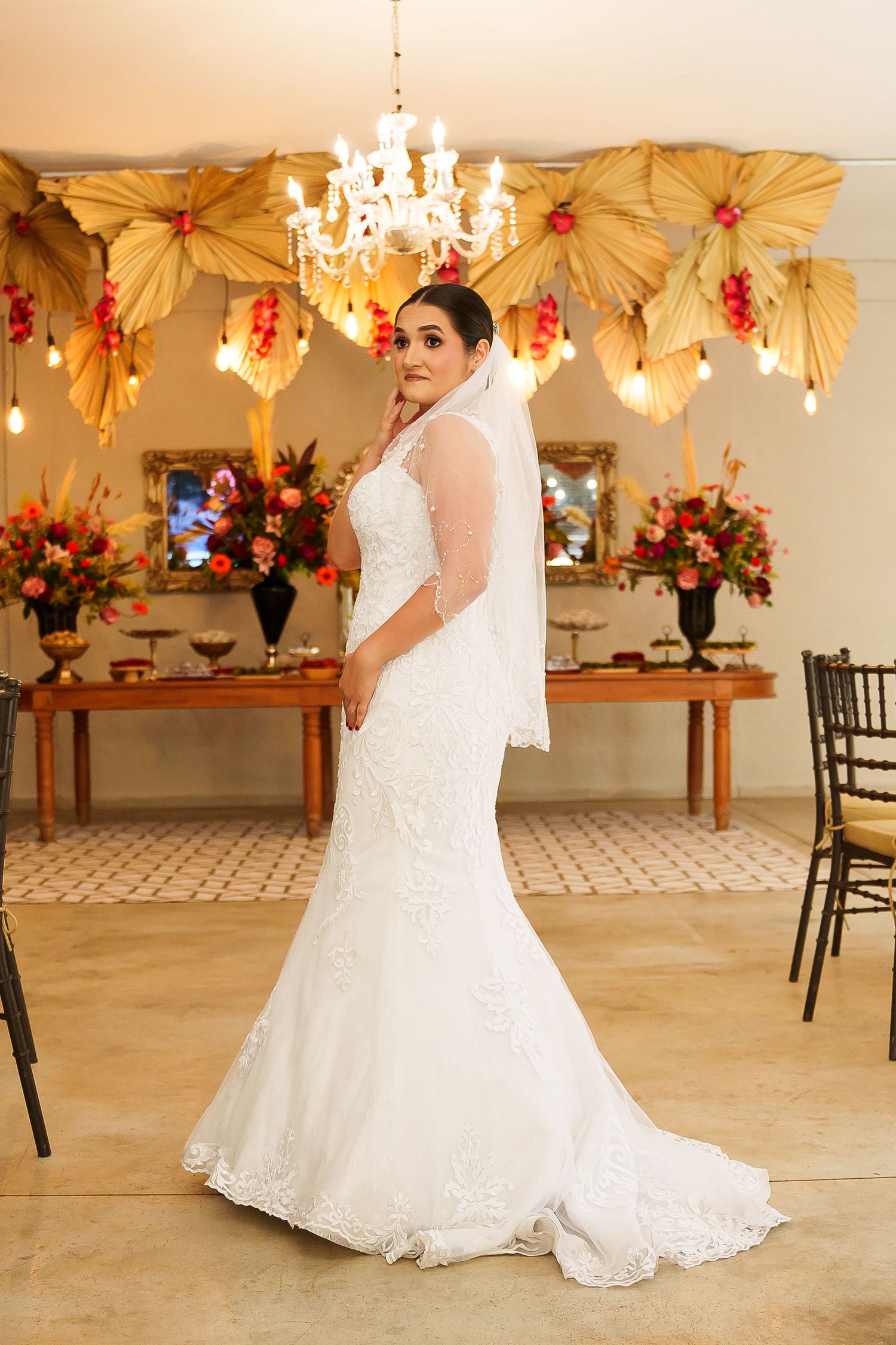 Bride in white lace mermaid wedding gown with veil poses in decorated venue with floral arrangements and chandelier.