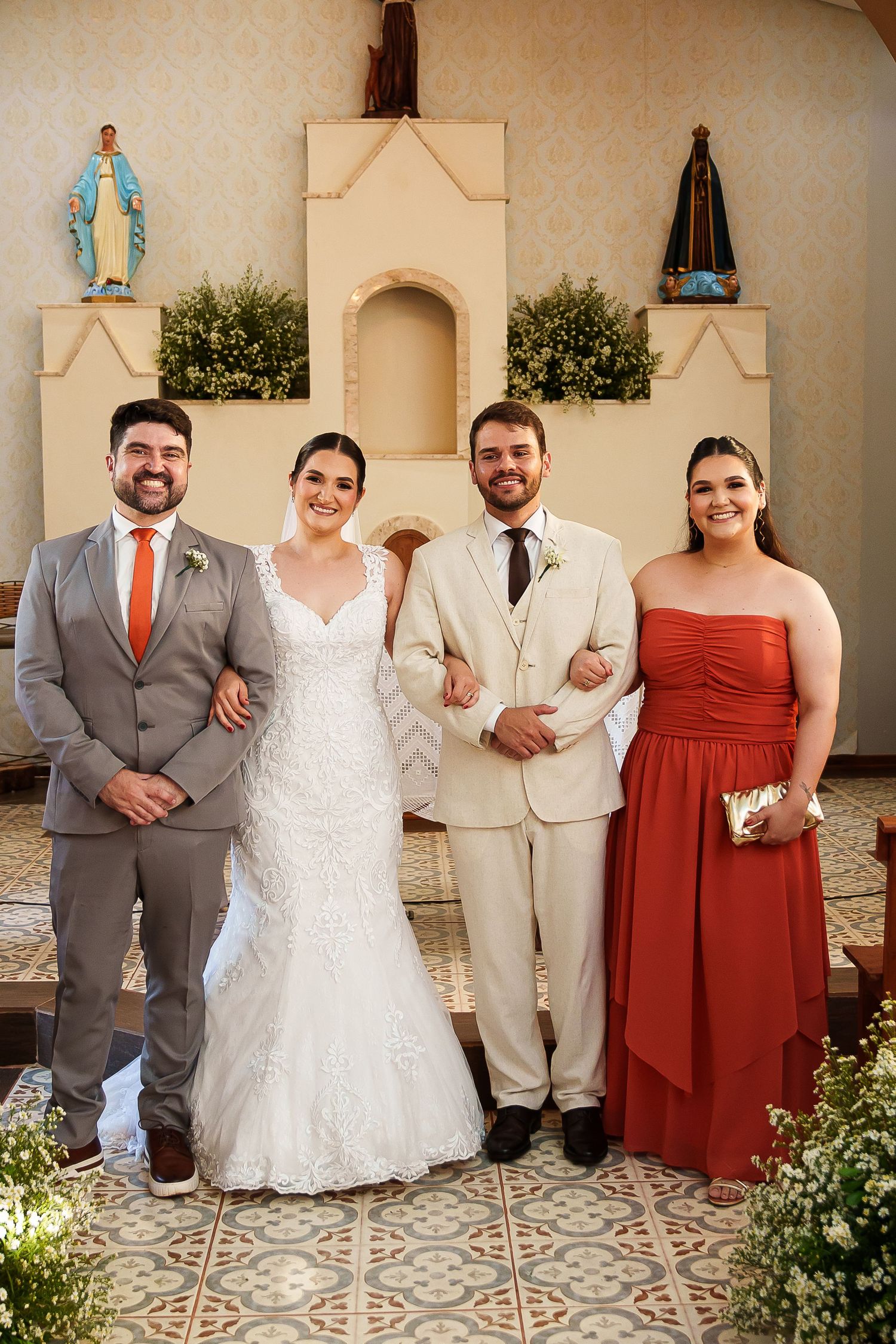 Wedding party posing in church with bride in white gown, groom in cream suit, groomsman in gray, and bridesmaid in red dress.