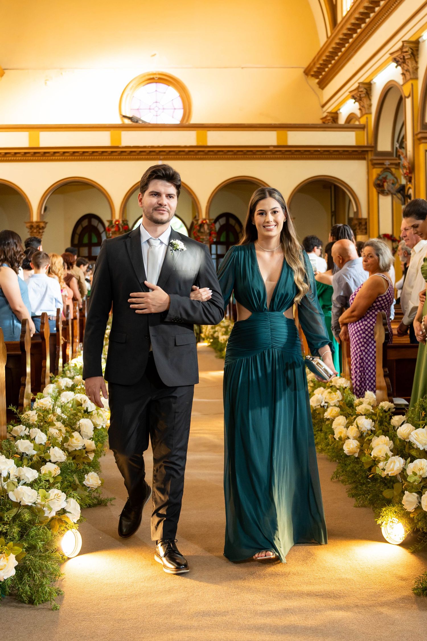 Groomsman in black suit escorts bridesmaid in emerald green gown down flower-lined church aisle.
