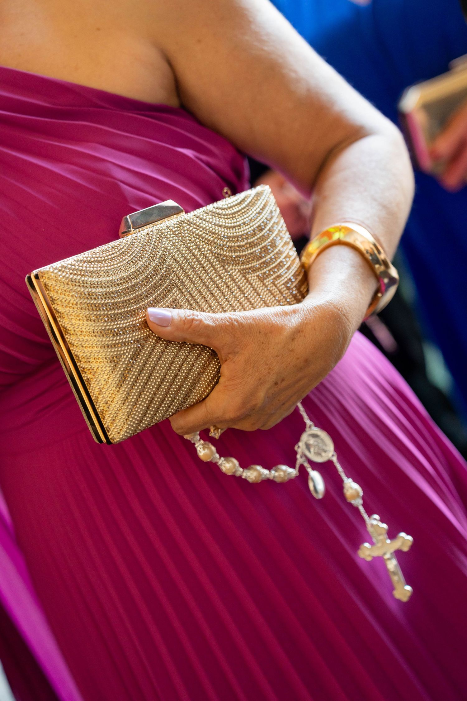 Woman in magenta gown holding gold clutch purse and pearl rosary with cross, wearing gold bracelet.