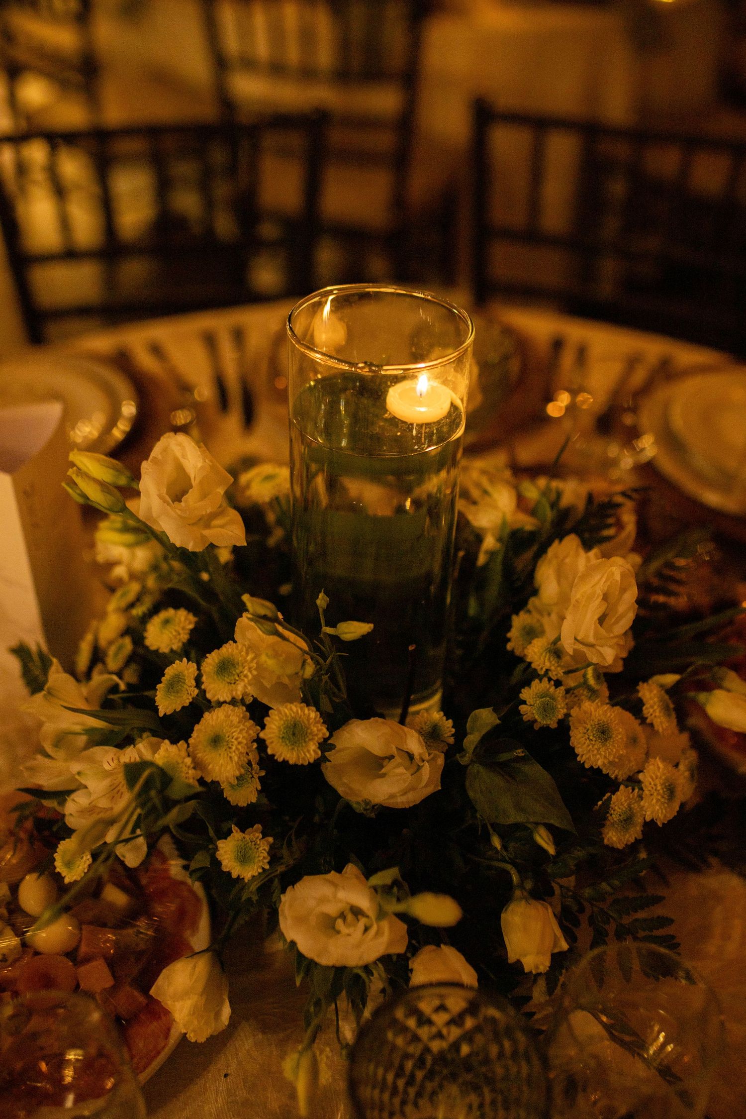 Elegant wedding centerpiece with white flowers and a glowing candle in a glass cylinder vase on a round table.