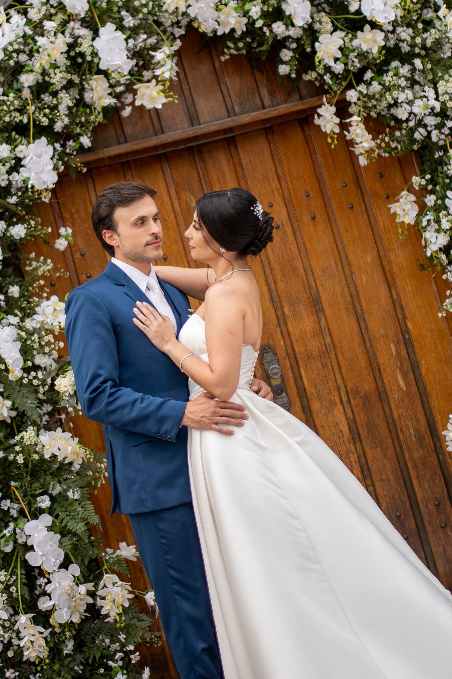 Bride and groom embrace under a floral arch with white flowers and wooden door backdrop on their wedding day.