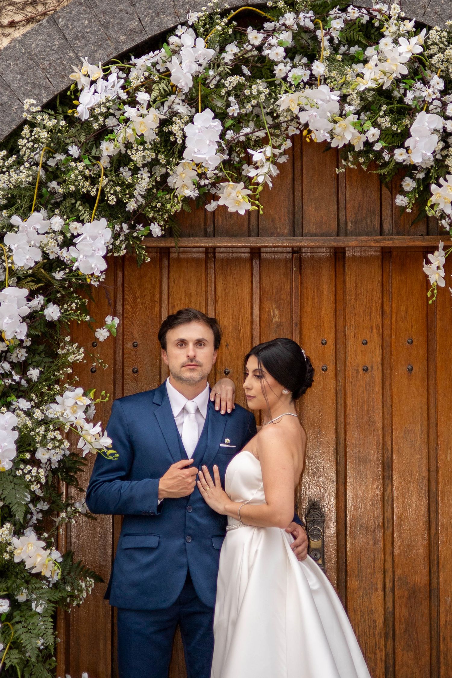 Bride and groom posing under a floral arch with white flowers in front of a wooden door.
