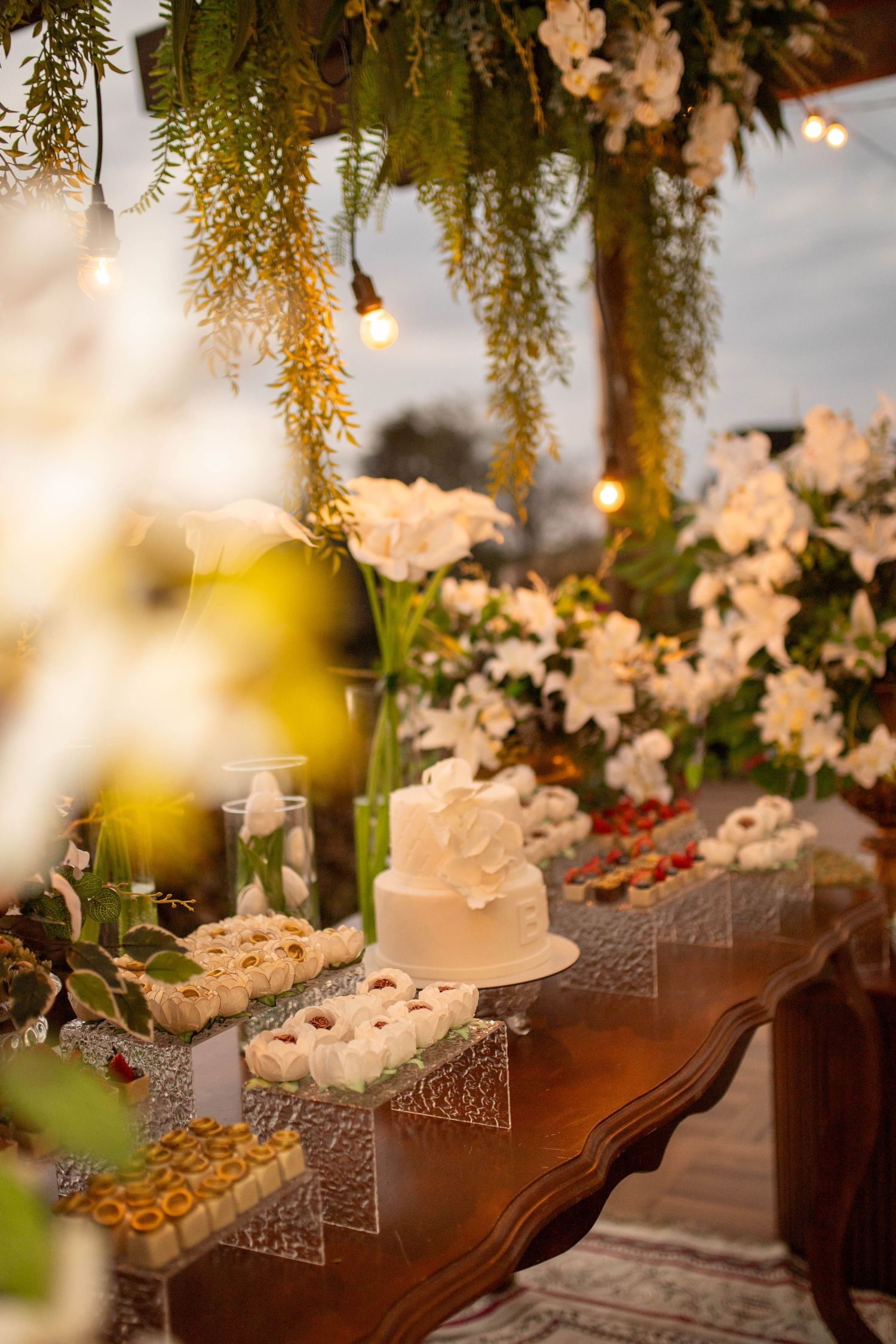 Elegant wedding dessert table with white tiered cake, assorted sweets, white floral arrangements, and warm string lights overhead.