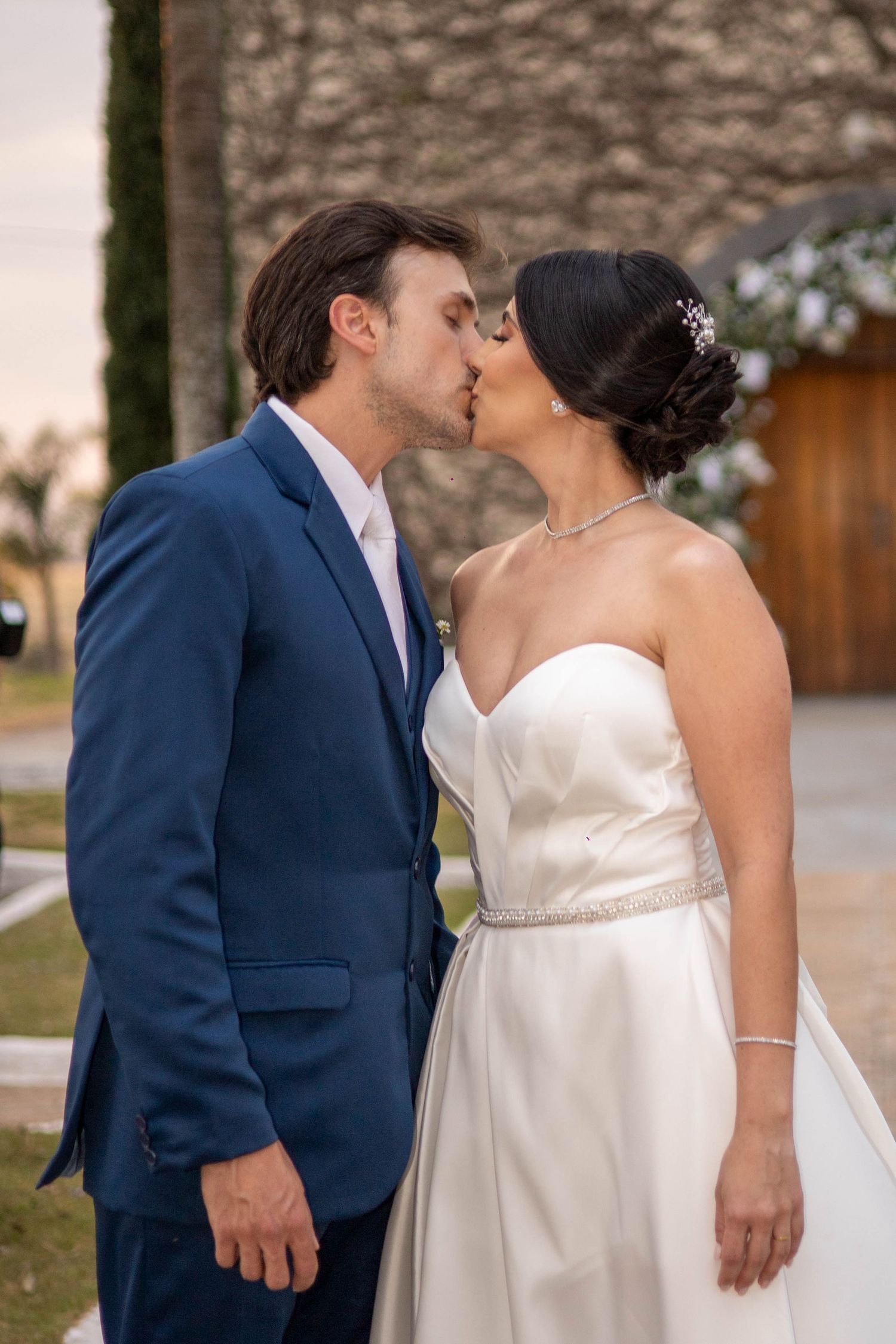 A bride and groom share a romantic kiss outdoors, she in a white strapless gown and he in a blue suit.