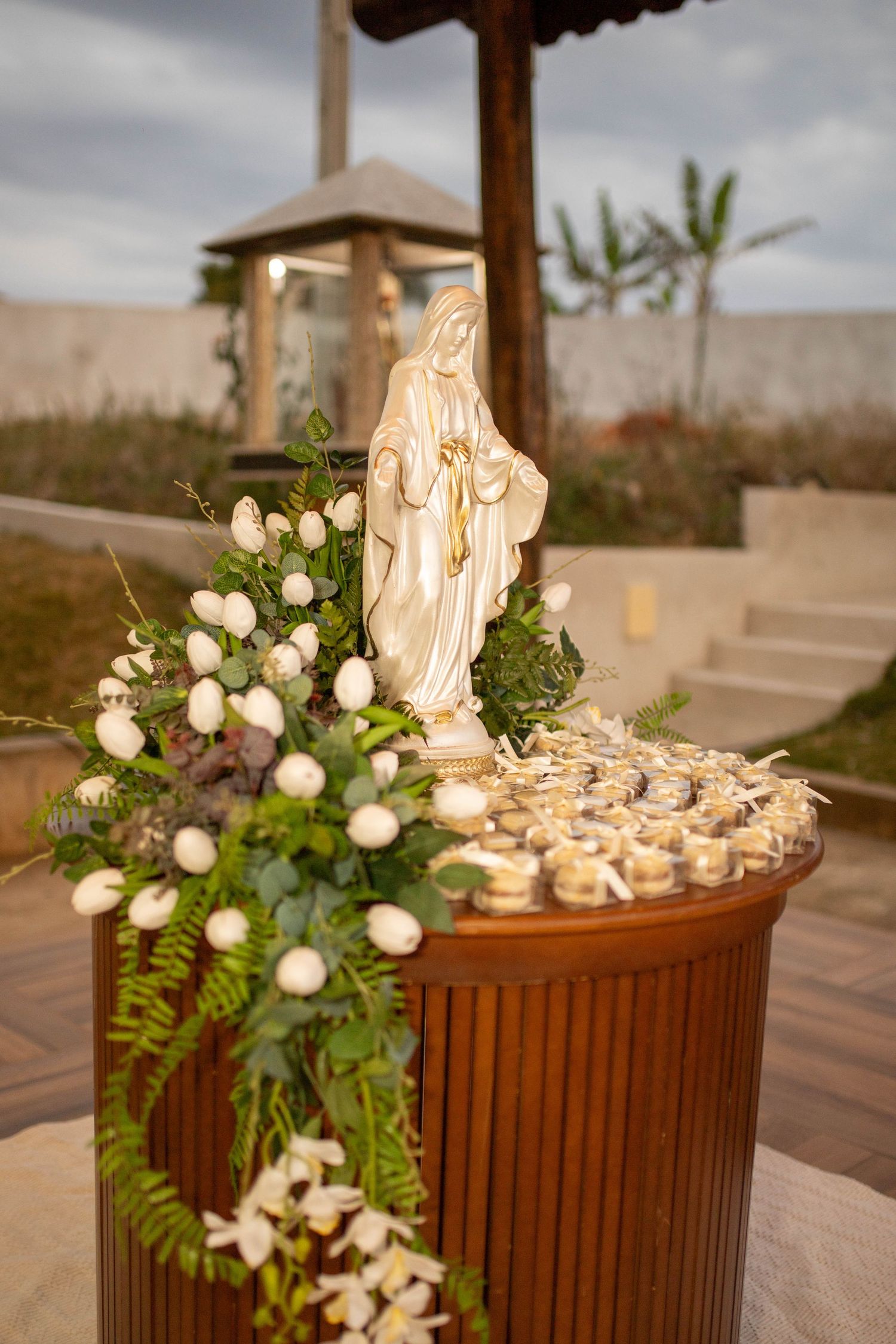White Virgin Mary statue surrounded by white floral arrangements on wooden table at outdoor religious ceremony.