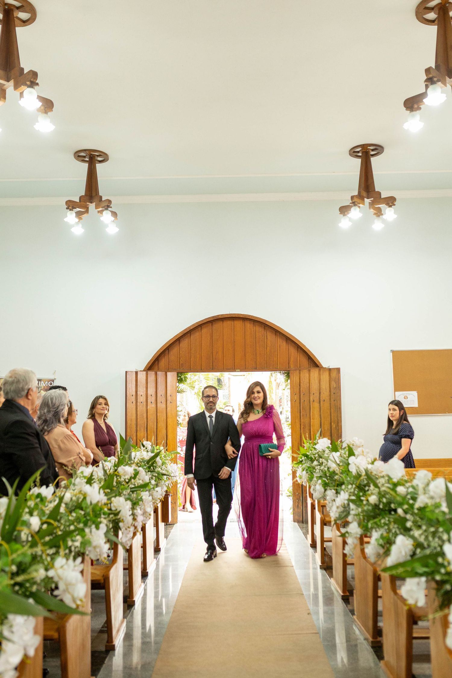 Couple walks down a flower-adorned church aisle, woman in magenta gown, man in dark suit, guests seated on wooden pews.