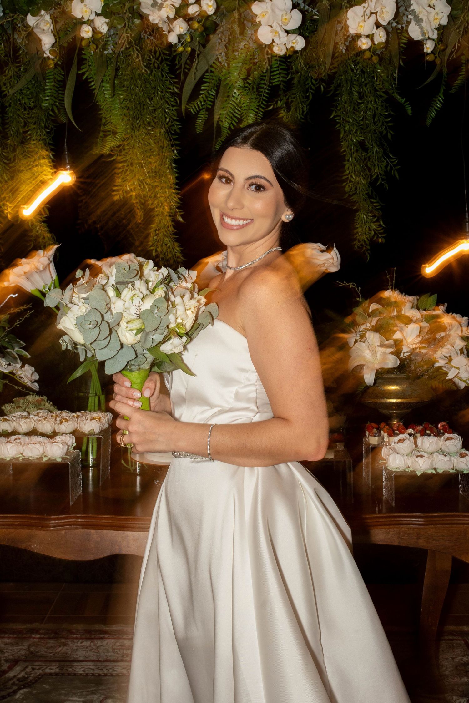 Smiling bride in strapless white satin gown holding white floral bouquet with greenery at elegant wedding reception.