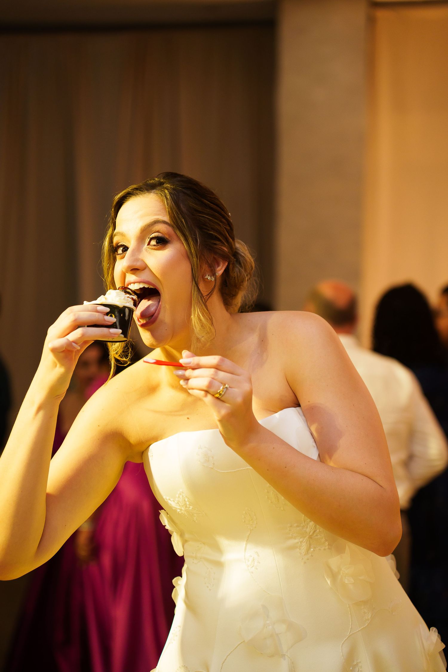 Bride in white strapless gown joyfully eating a cupcake at her wedding reception.