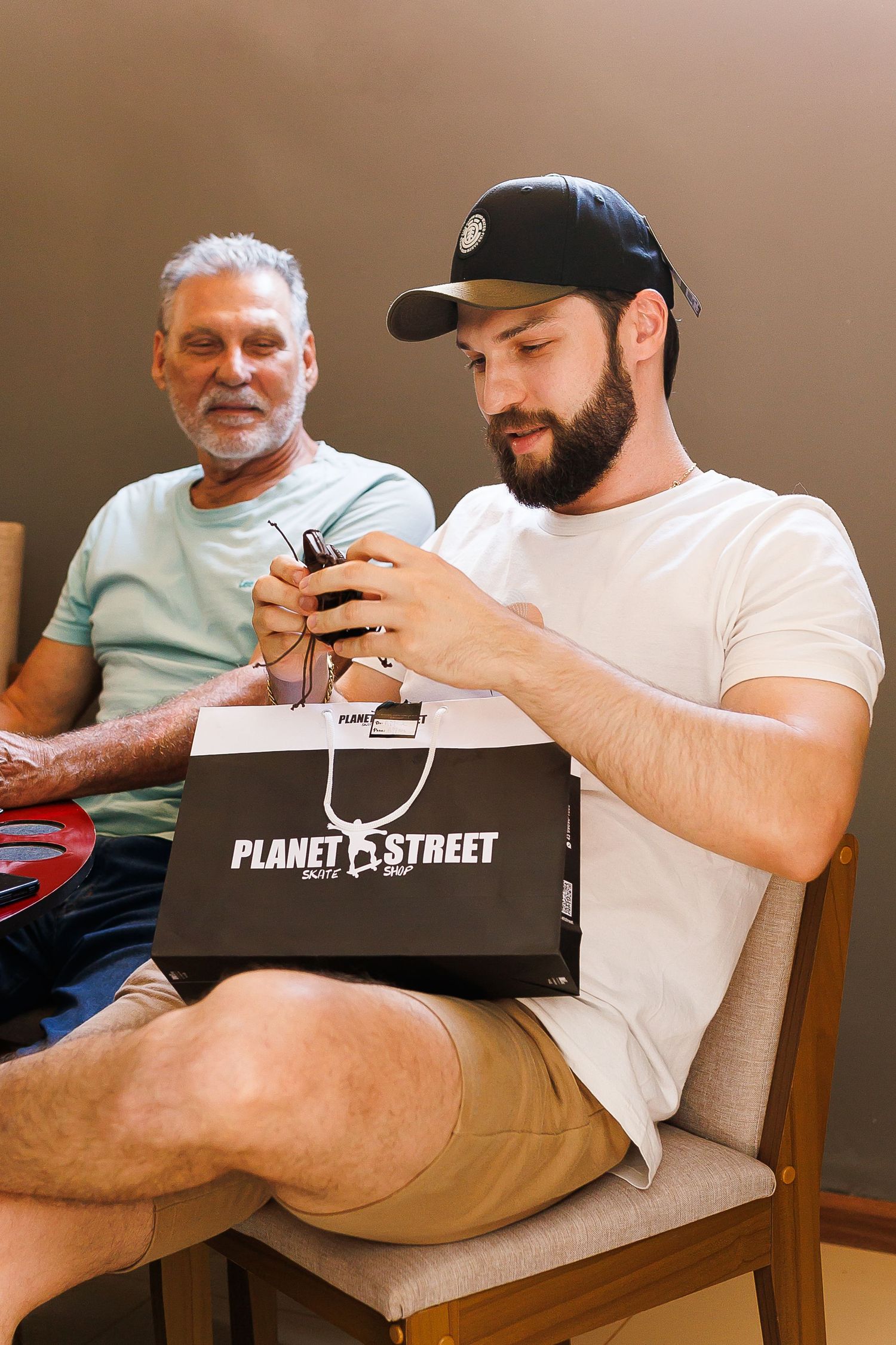 Young bearded man in cap examines Planet Street shopping bag while older man sits smiling in background.