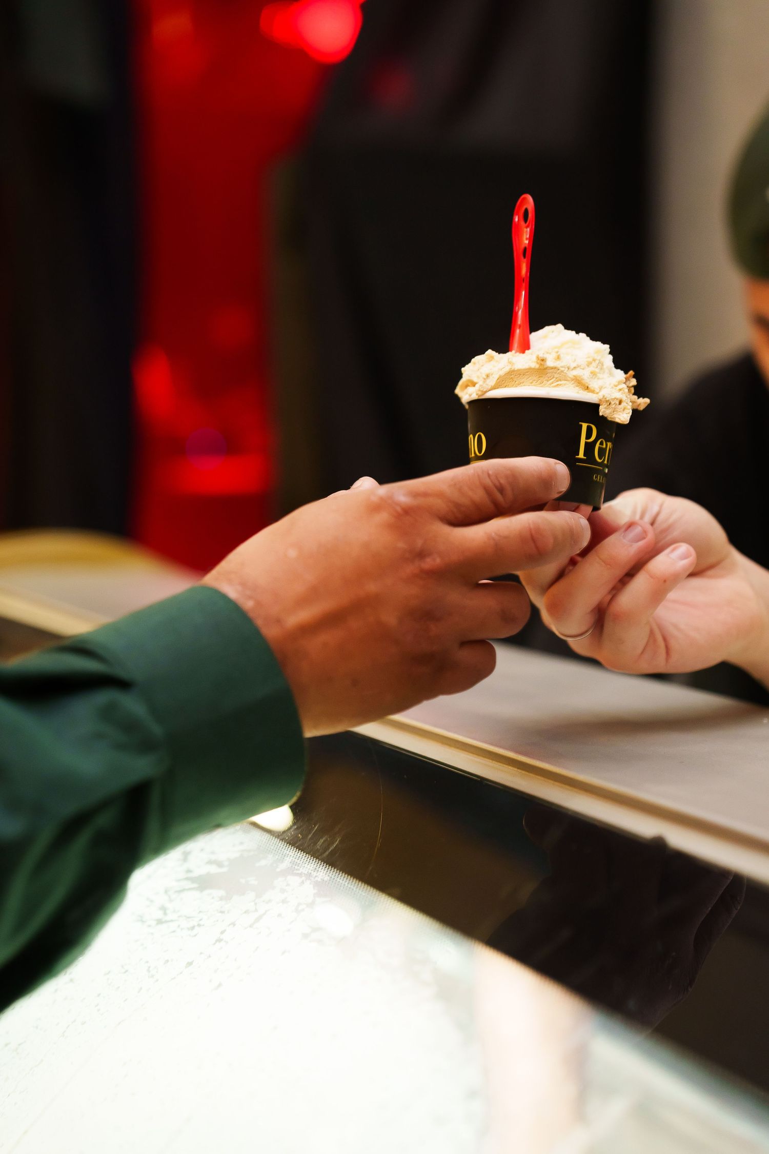 Two hands exchanging a black cup of ice cream topped with whipped cream and a red spoon at a counter.