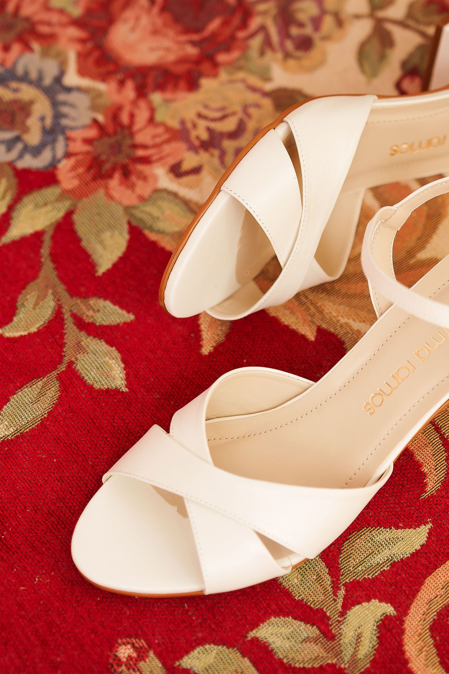 White strappy heeled sandals with crossover toe straps displayed on a red floral rug.