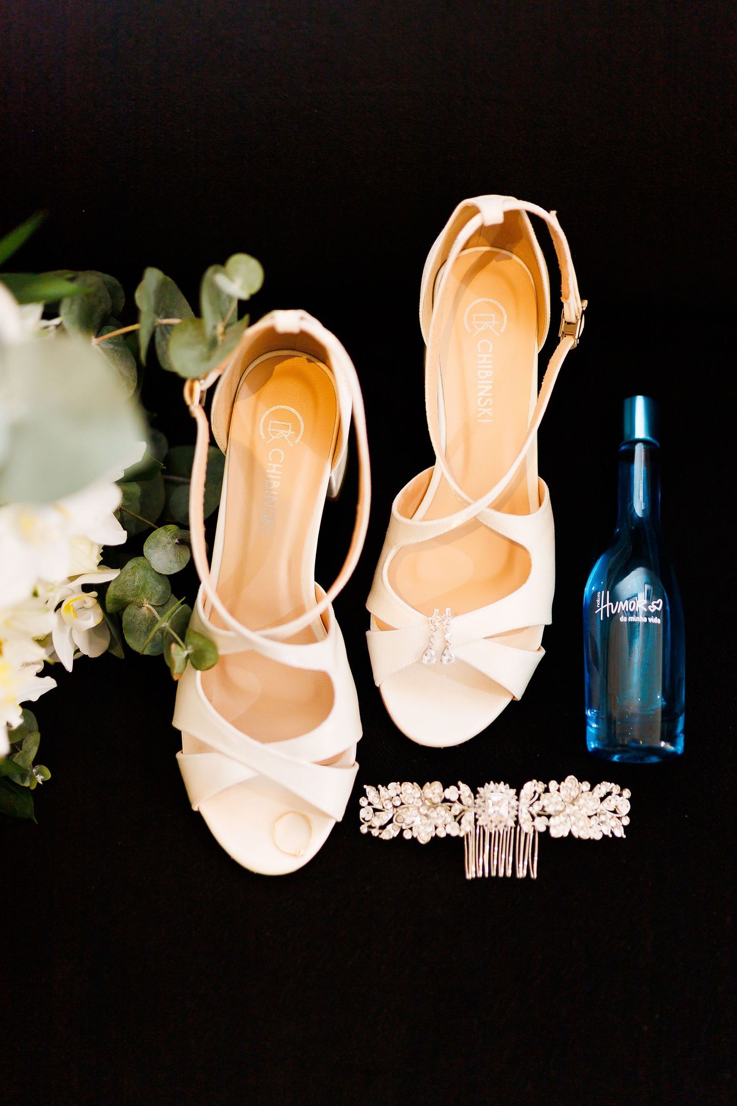 Elegant bridal flat lay featuring white strappy heels, crystal hair comb, blue perfume bottle, and white flowers on black background.
