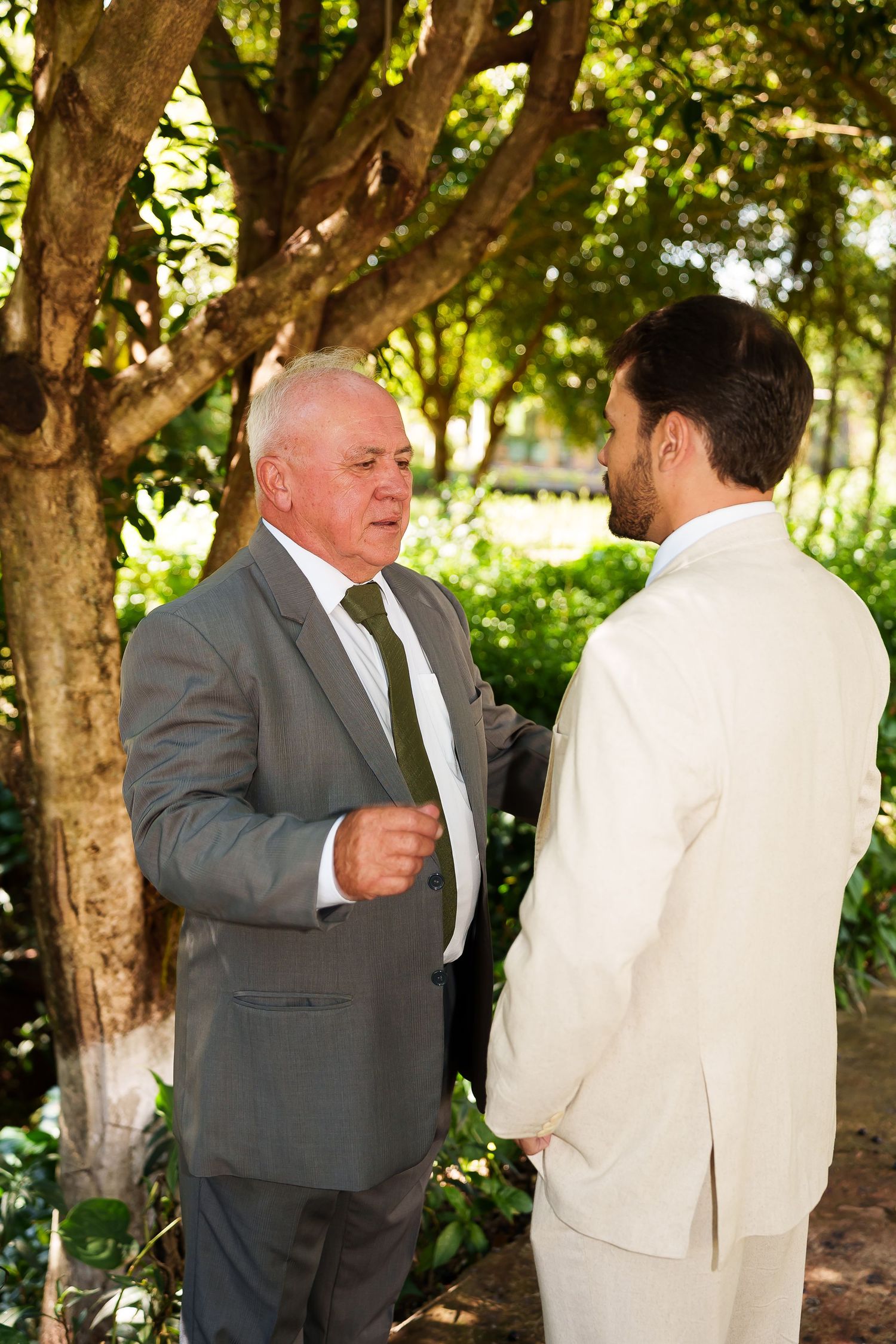 Two men in suits conversing outdoors under green trees, possibly at a wedding ceremony.