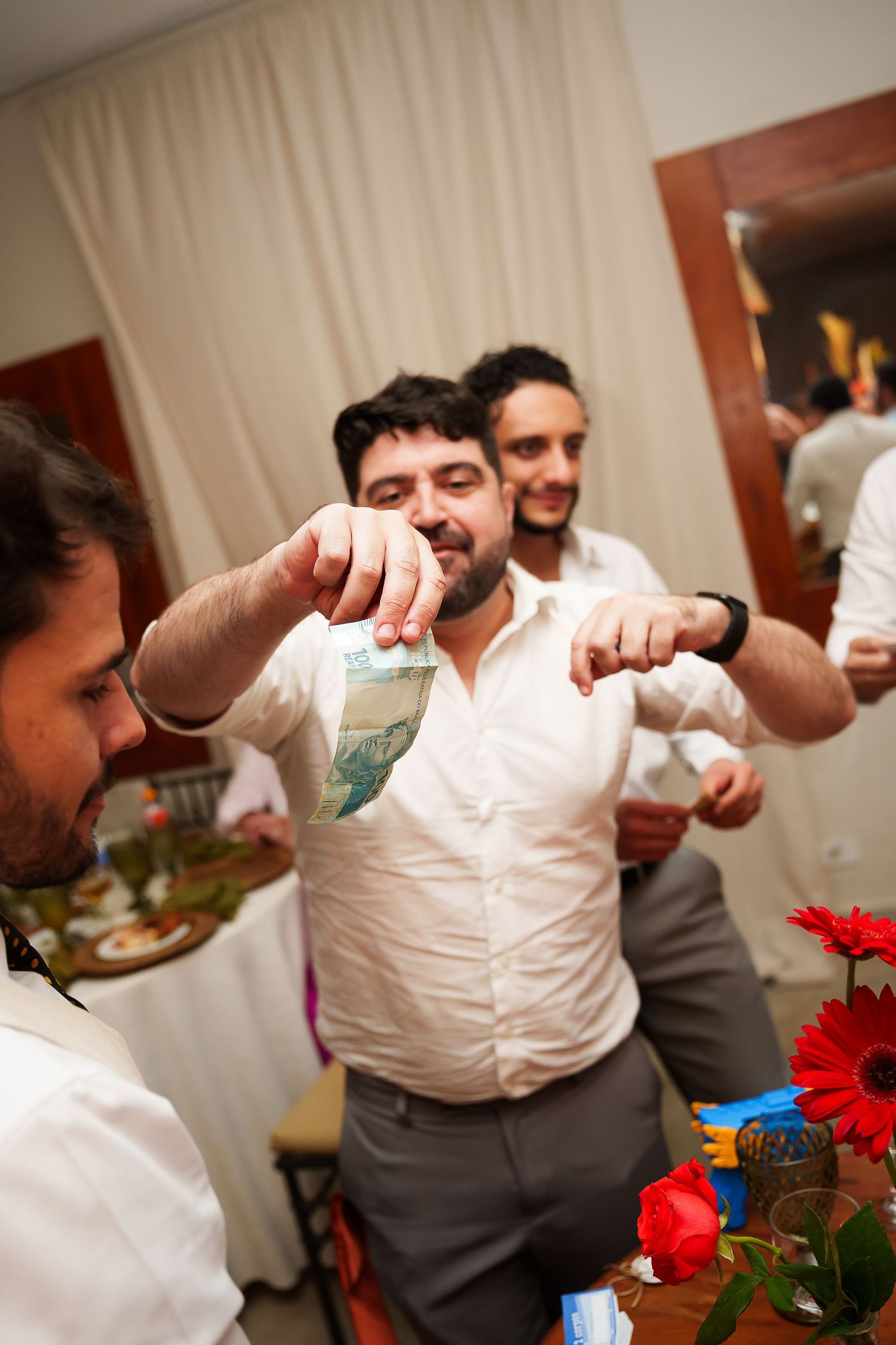 Men in white shirts at a party, one holding currency bills while others smile in a decorated event venue.