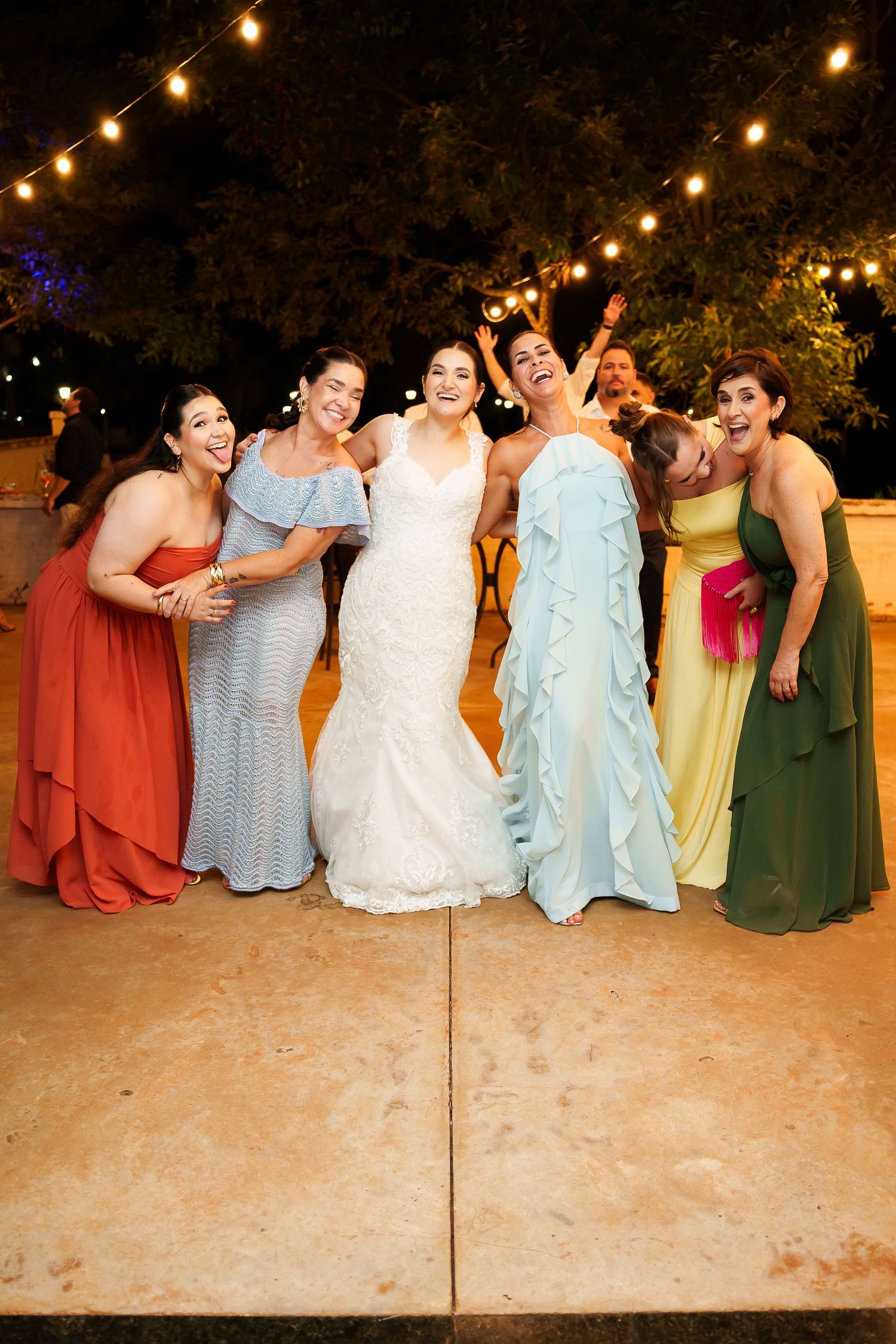 Bride in white gown poses with five bridesmaids in colorful dresses at an outdoor evening wedding reception.