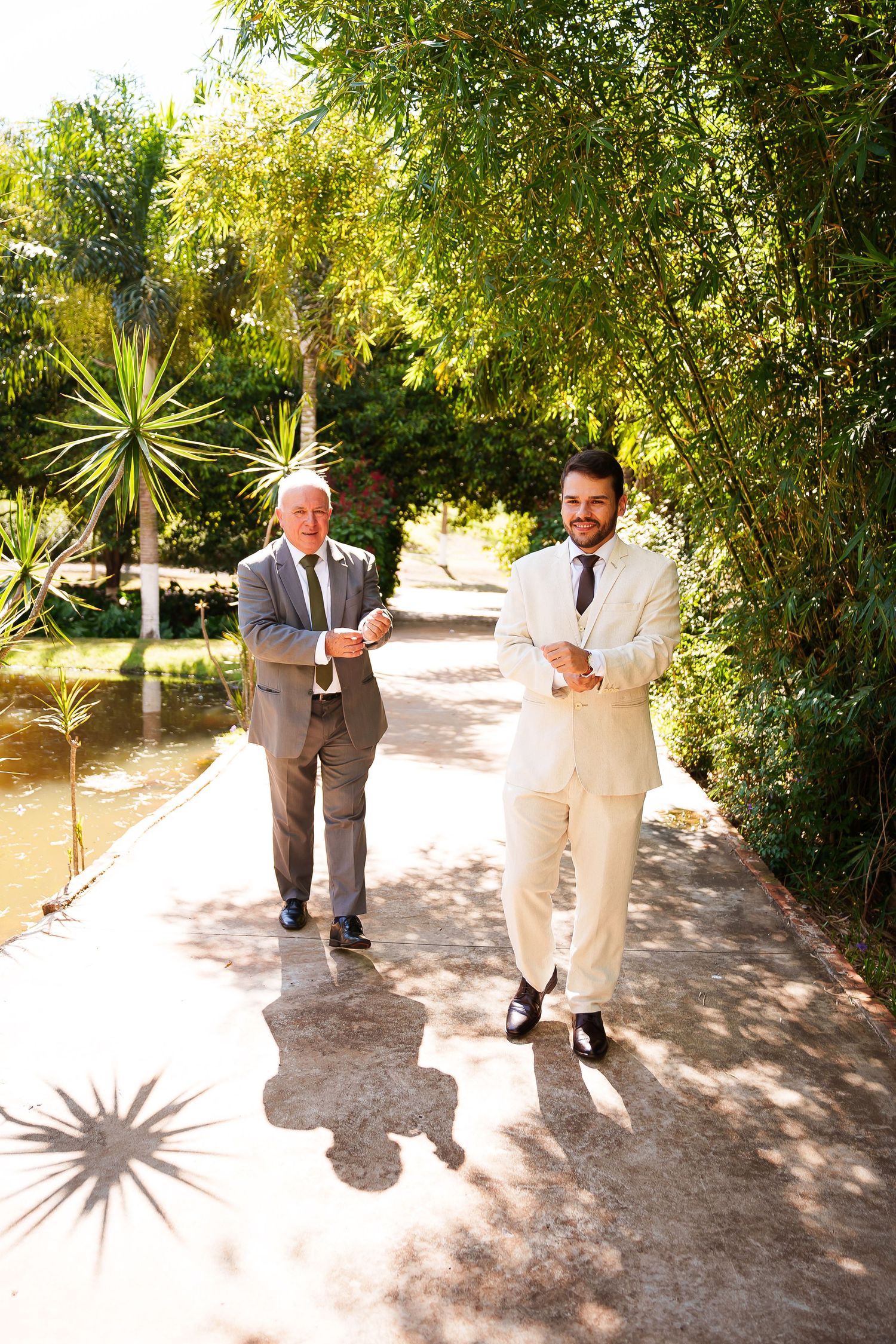 Two well-dressed men in suits walking along a sunlit garden path surrounded by lush green tropical plants.
