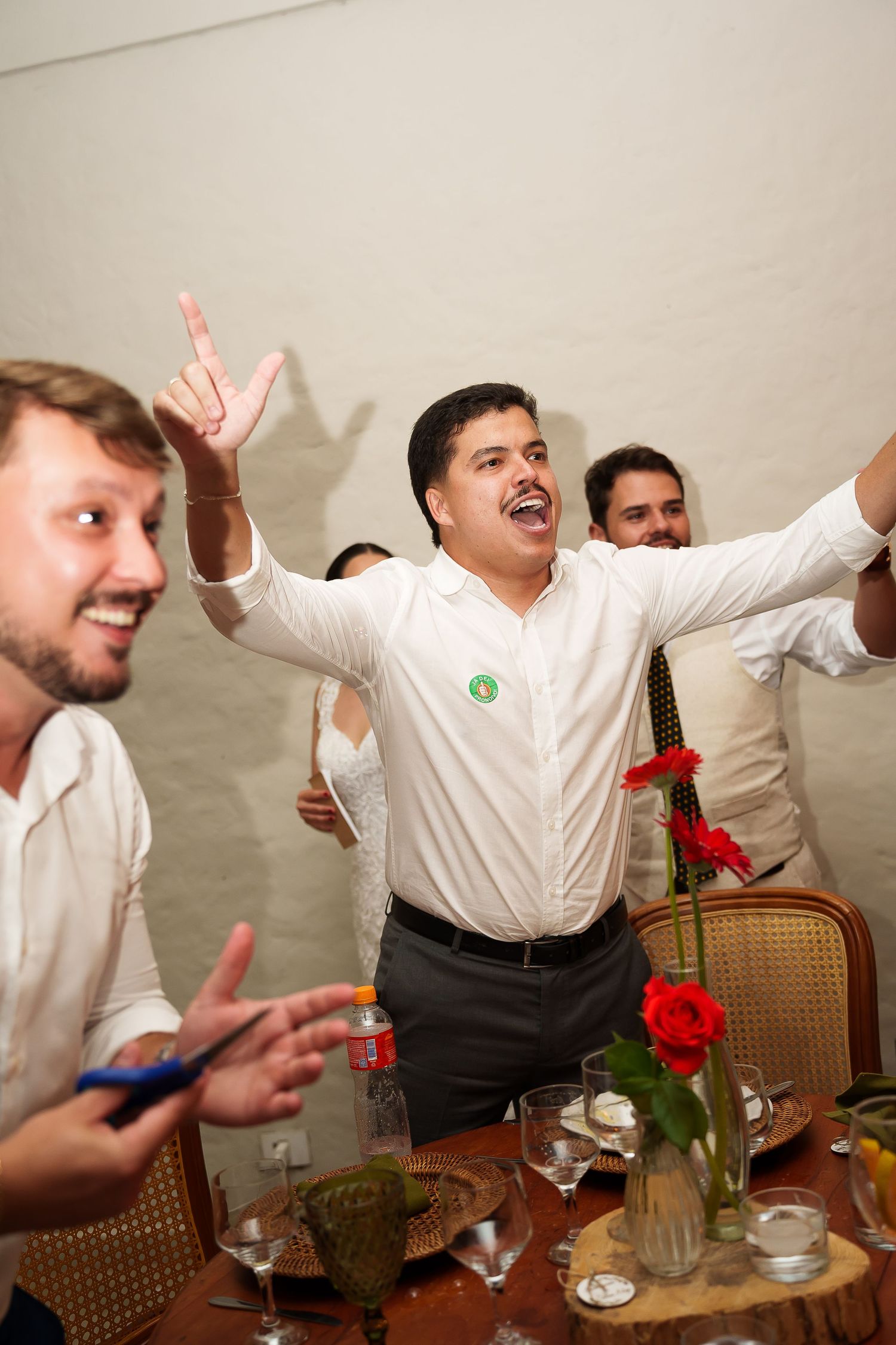 Man in white shirt celebrating with arms raised at a dinner party with friends and red flowers.
