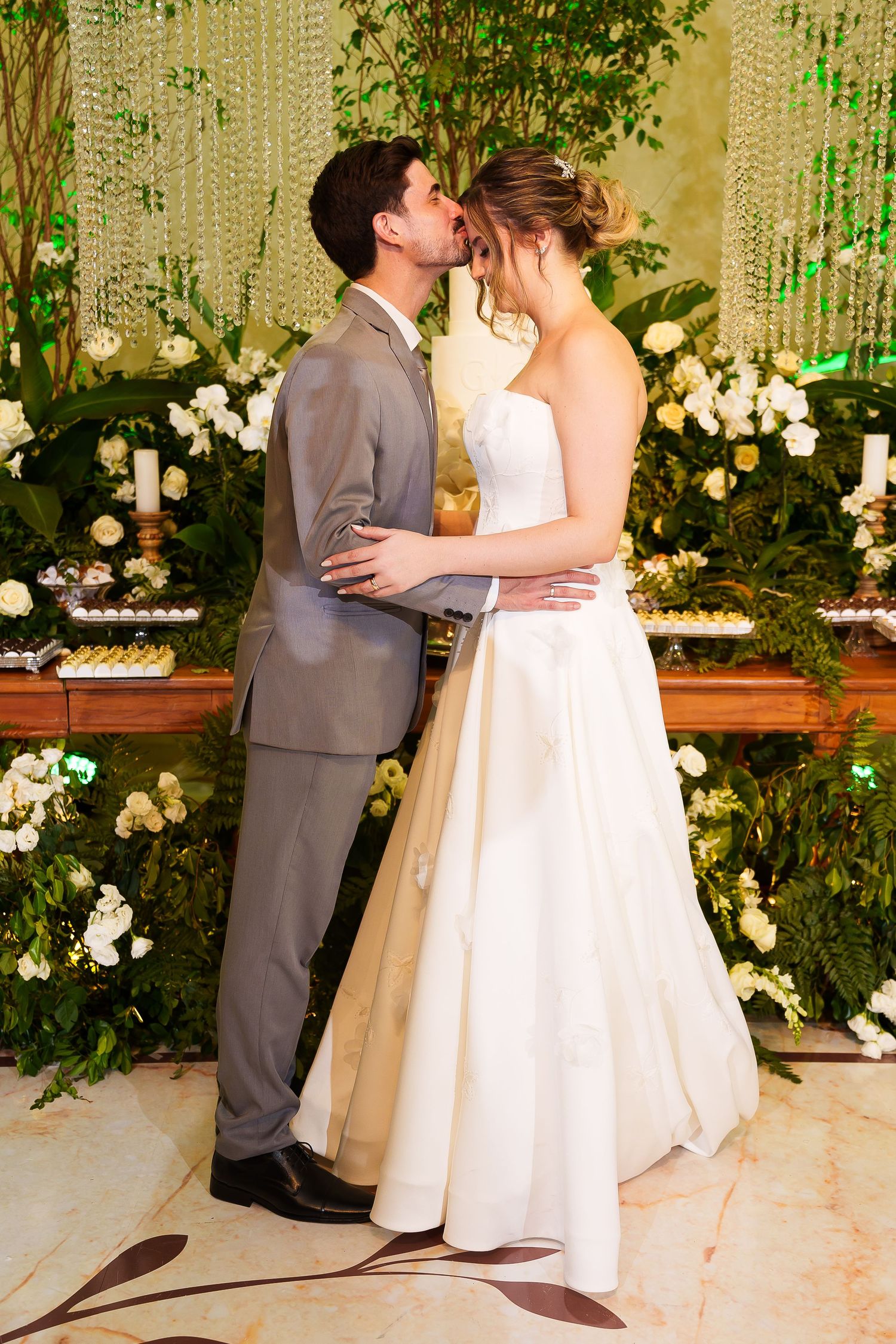 Bride and groom share a romantic kiss at their wedding reception surrounded by lush greenery and white floral arrangements.