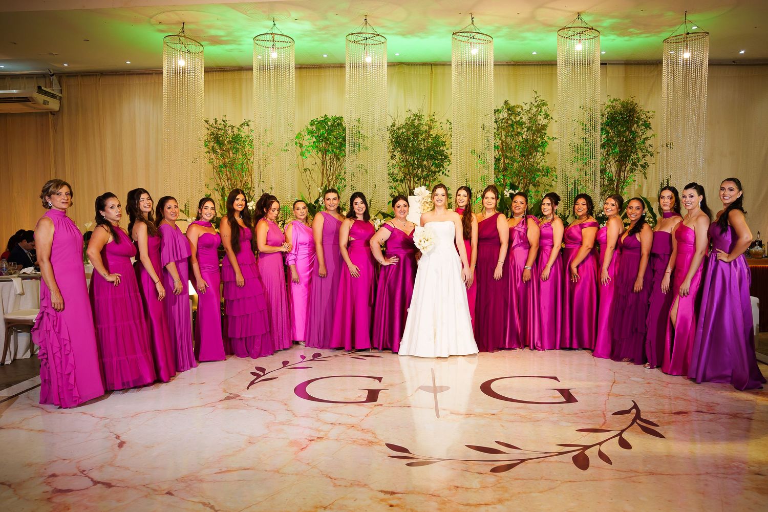 Bride in white gown poses with large bridal party in fuchsia gowns on monogrammed dance floor at elegant wedding reception.