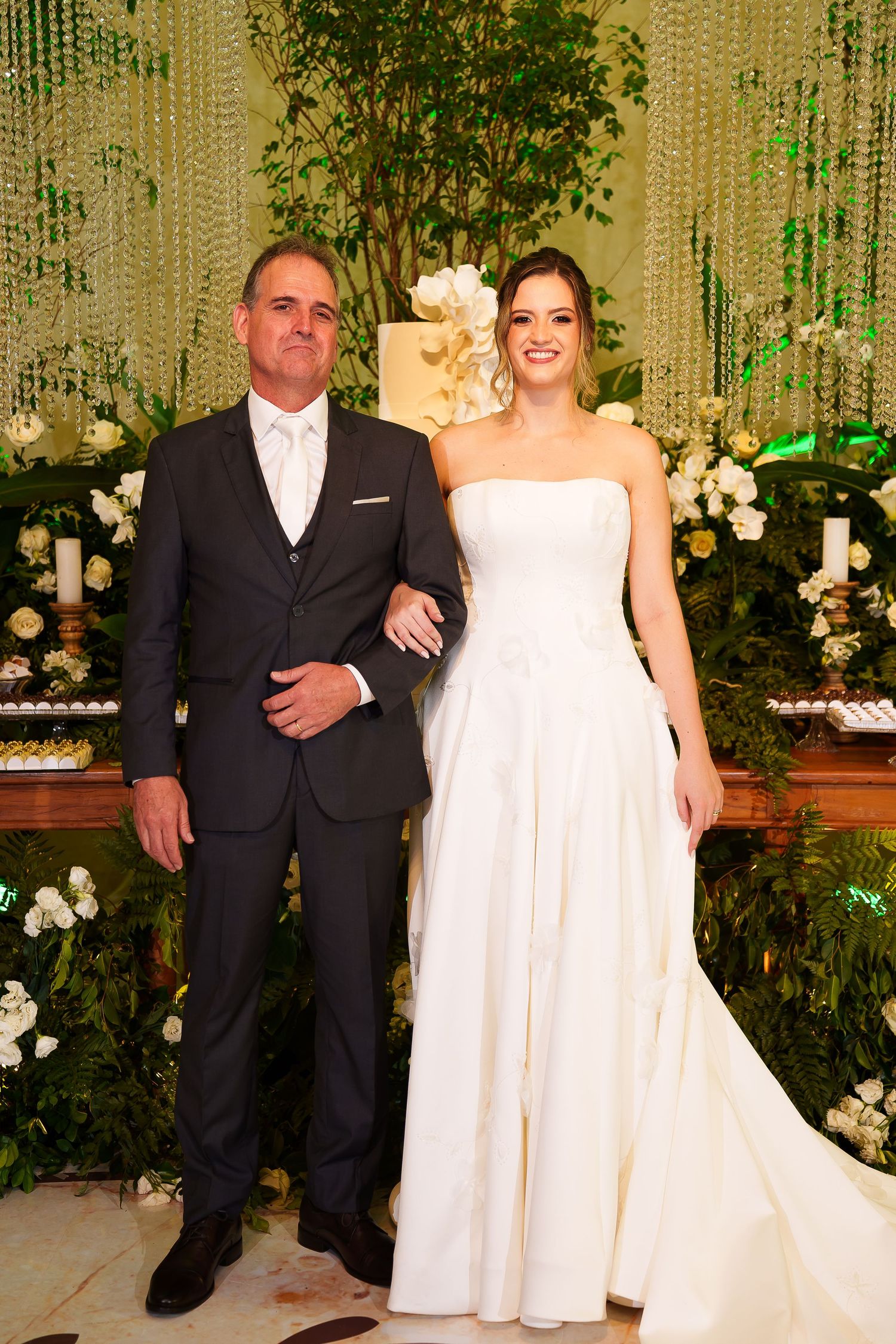 Bride in strapless white gown and groom in dark suit pose together at elegant wedding reception with floral decor.