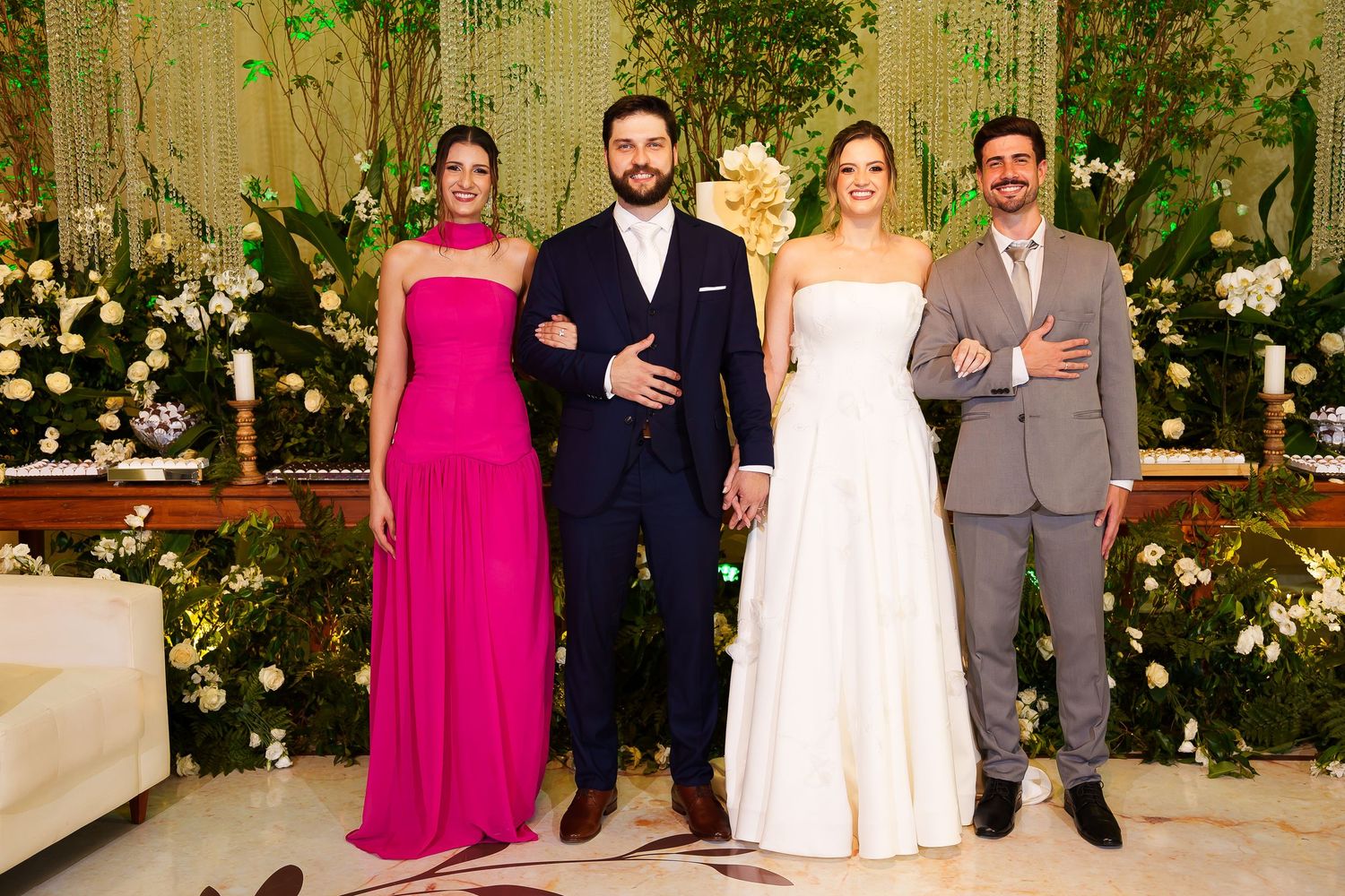 Two couples pose elegantly at a wedding reception, one in pink gown and one in white bridal dress, with floral decor.