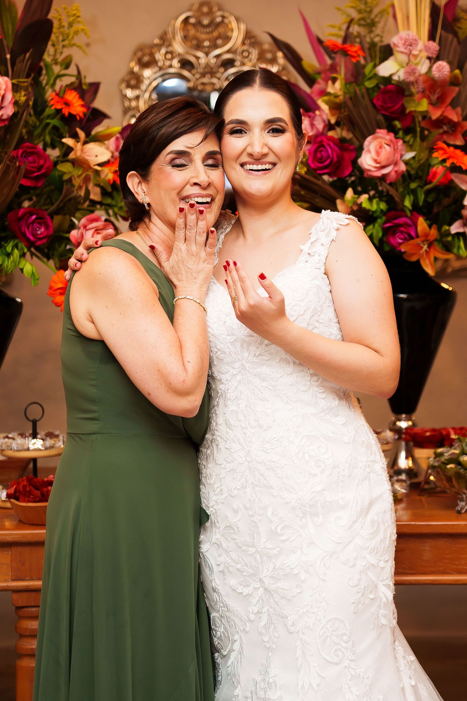 Bride in white lace gown and woman in green dress smile and show rings at floral wedding reception.