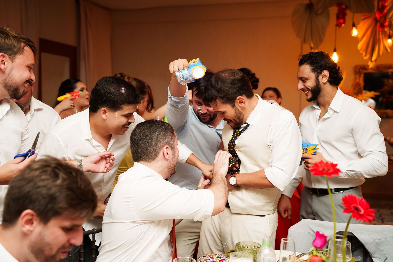 Guests in white shirts celebrate at a lively wedding reception, pouring drinks and laughing together.