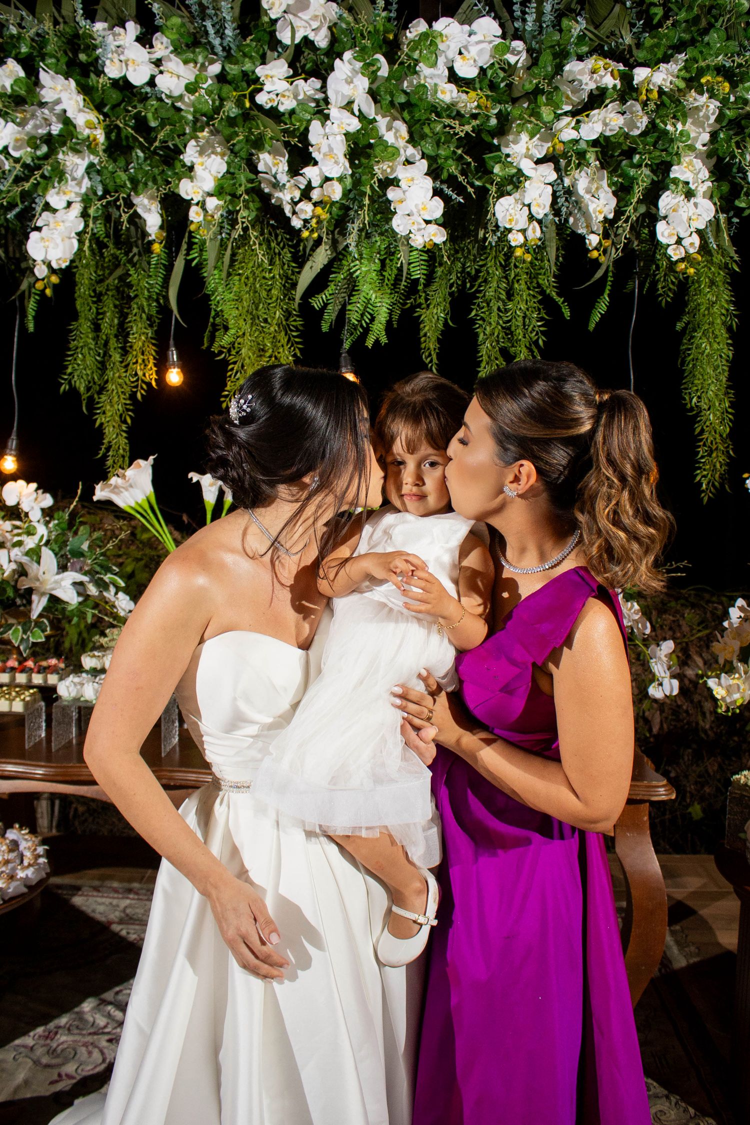 Bride in white gown and woman in purple dress kissing a child at a floral wedding reception.