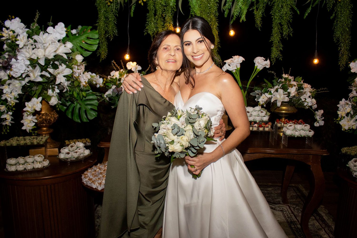 Bride in white gown holds bouquet beside older woman in olive dress, posing before floral wedding backdrop.