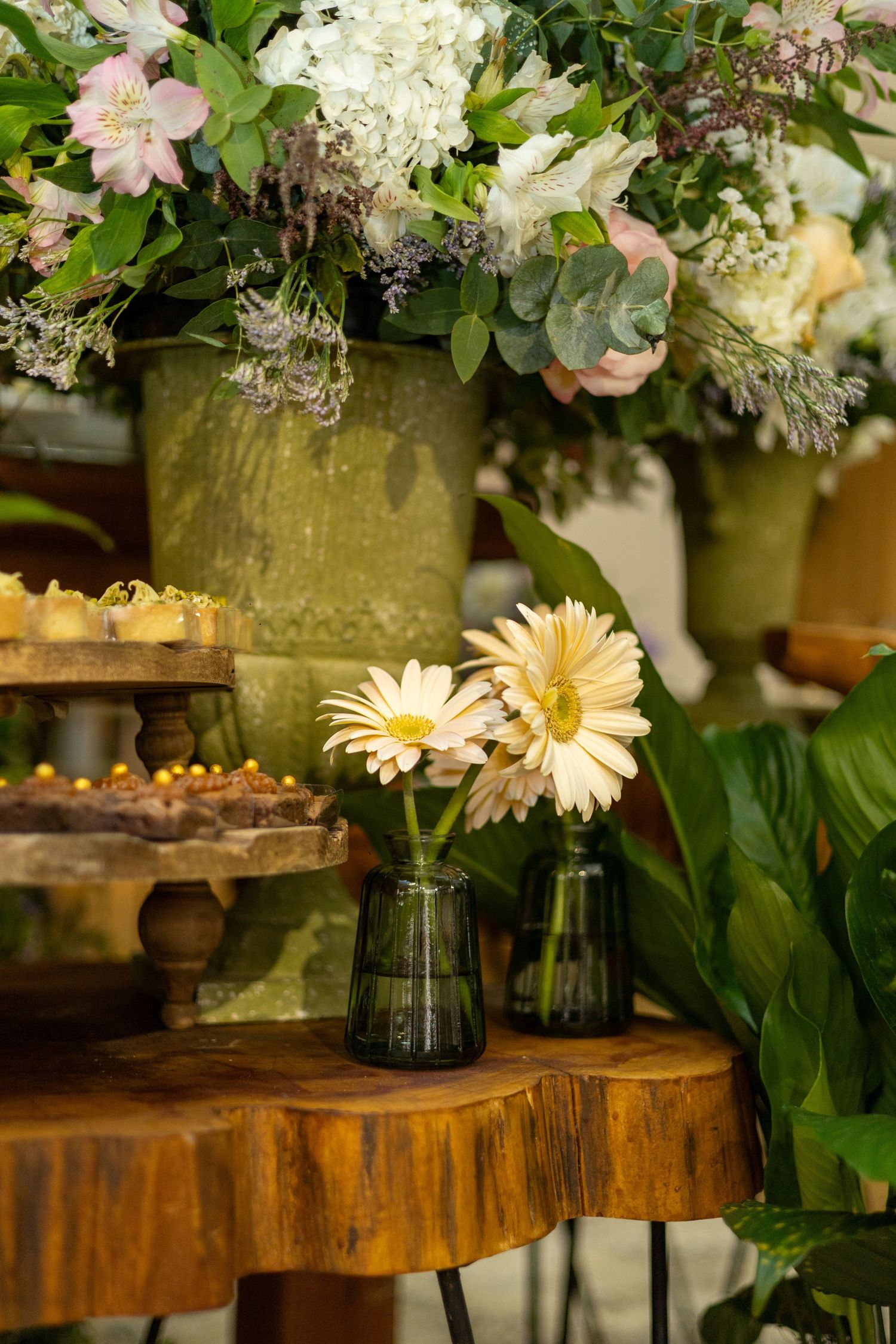 Yellow gerbera daisies in glass vases on rustic wood table with lush floral arrangements and dessert display.