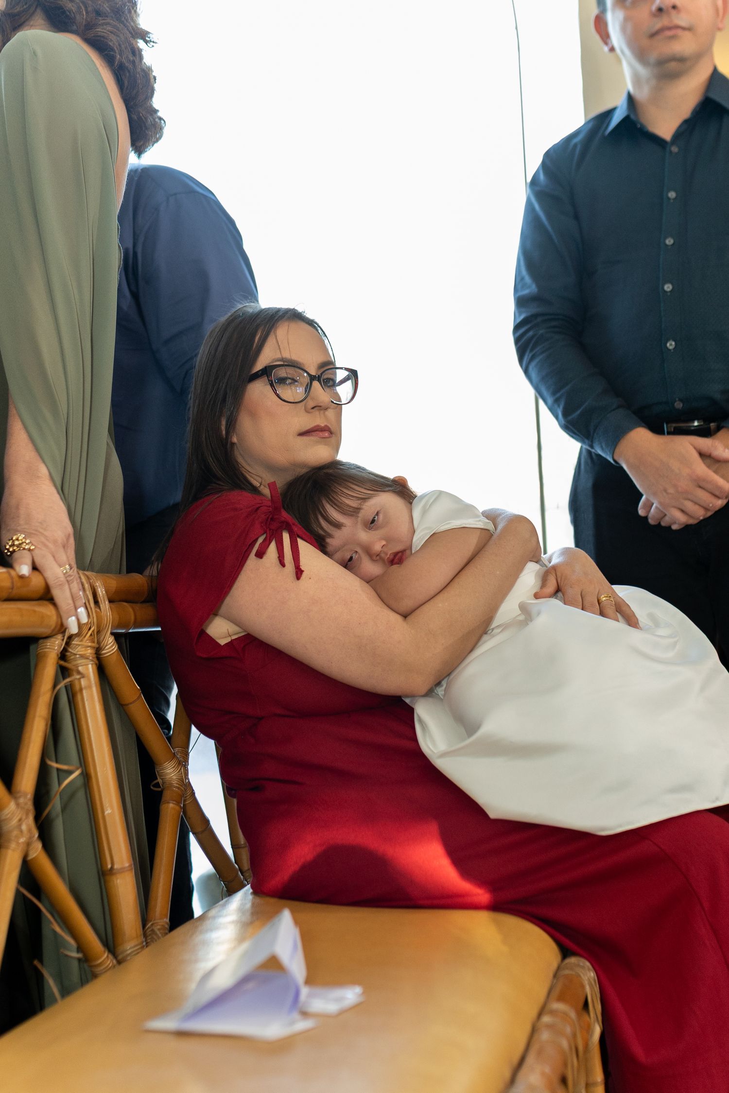Woman in red dress holding sleeping baby in white christening gown at a formal ceremony.