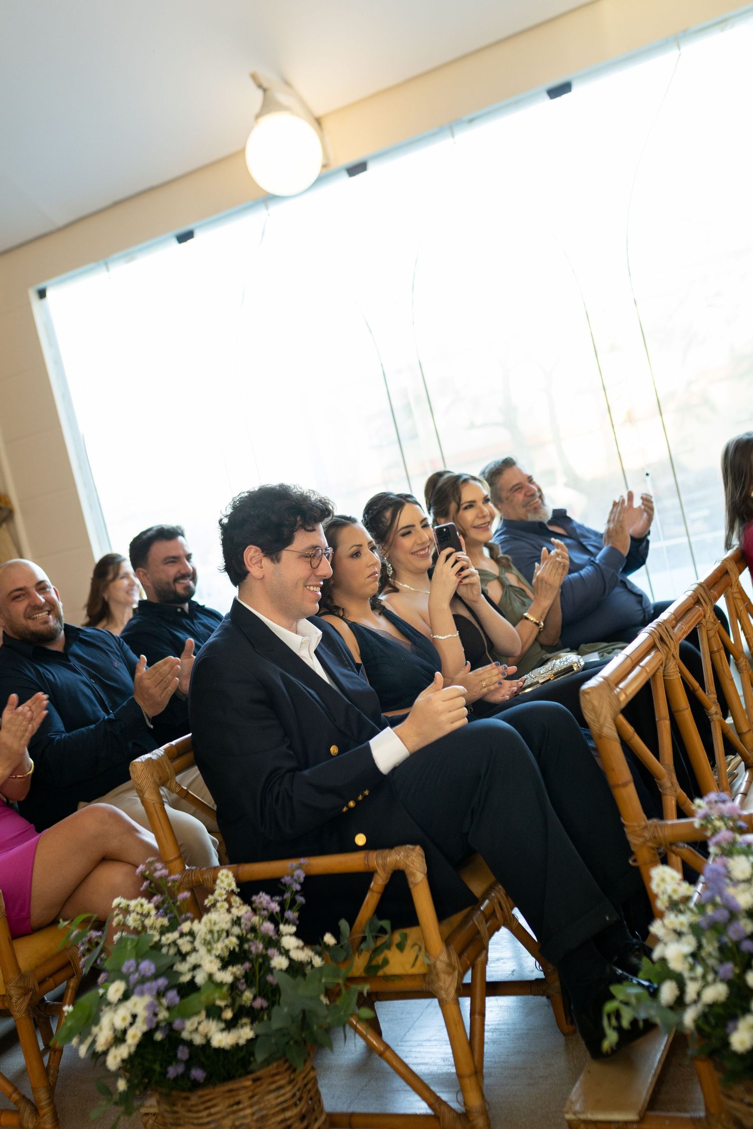Guests clapping and smiling during a wedding ceremony, seated on wooden chairs with floral arrangements in the foreground.