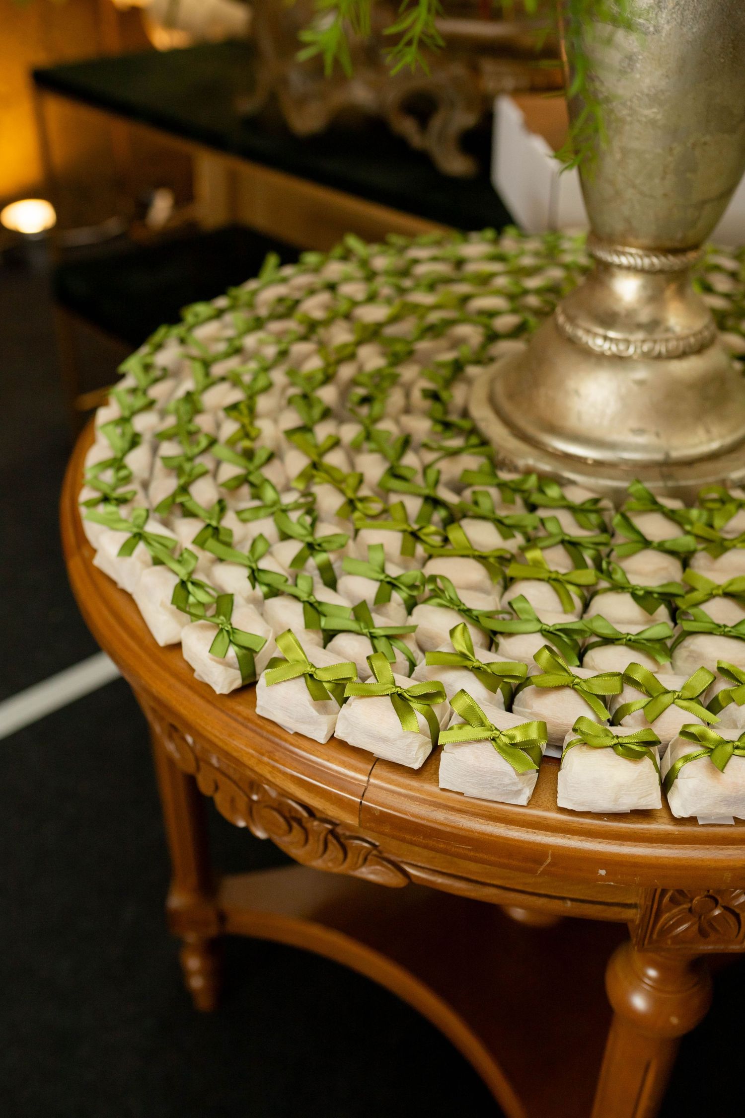 Elegant round wooden table displaying rows of white wedding favors tied with green satin bows, surrounding a silver vase centerpiece.
