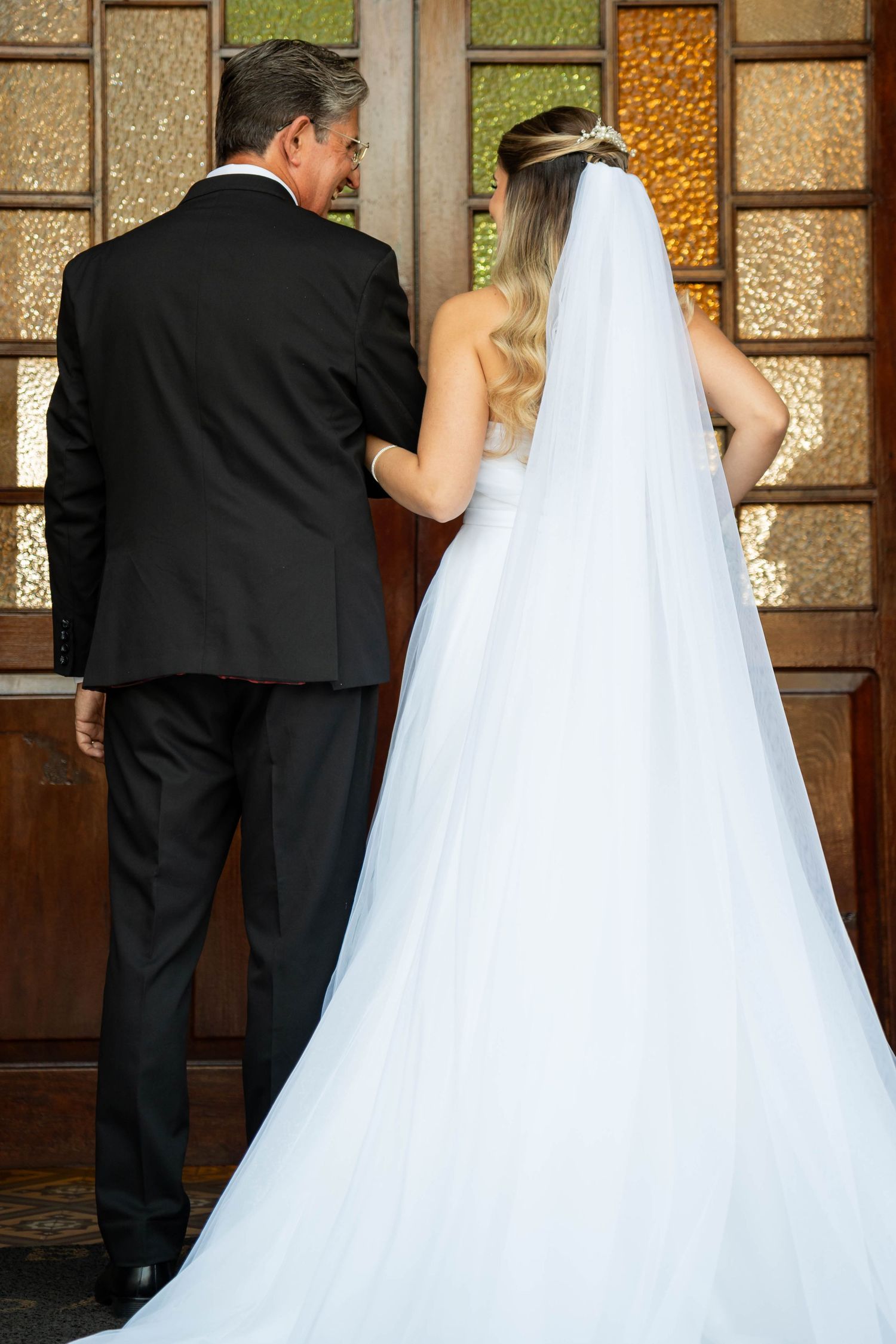 Bride in white gown and veil stands with man in black suit at ornate wooden church doors.