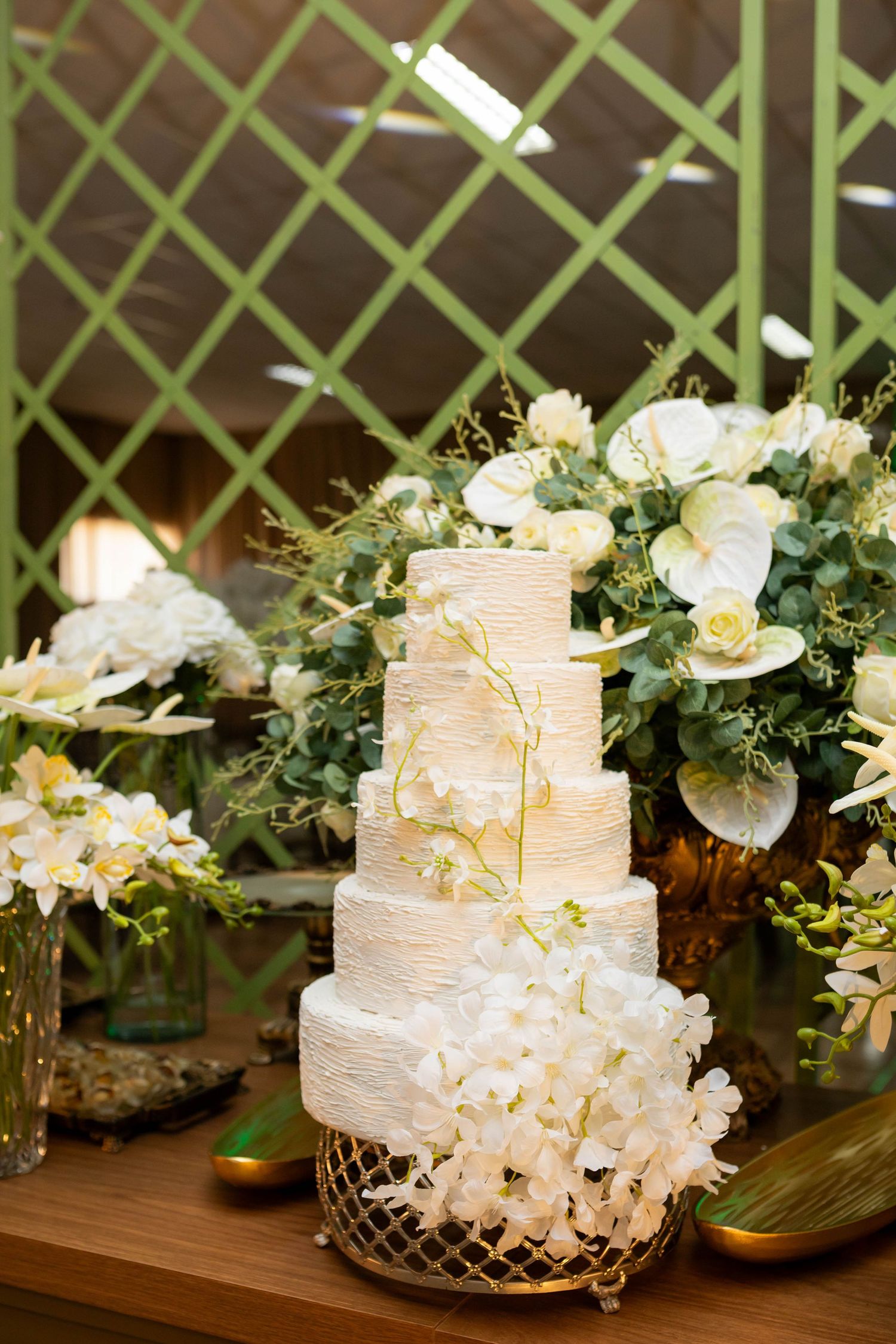 Elegant four-tier white wedding cake adorned with orchids on a gold stand, surrounded by lush floral arrangements.