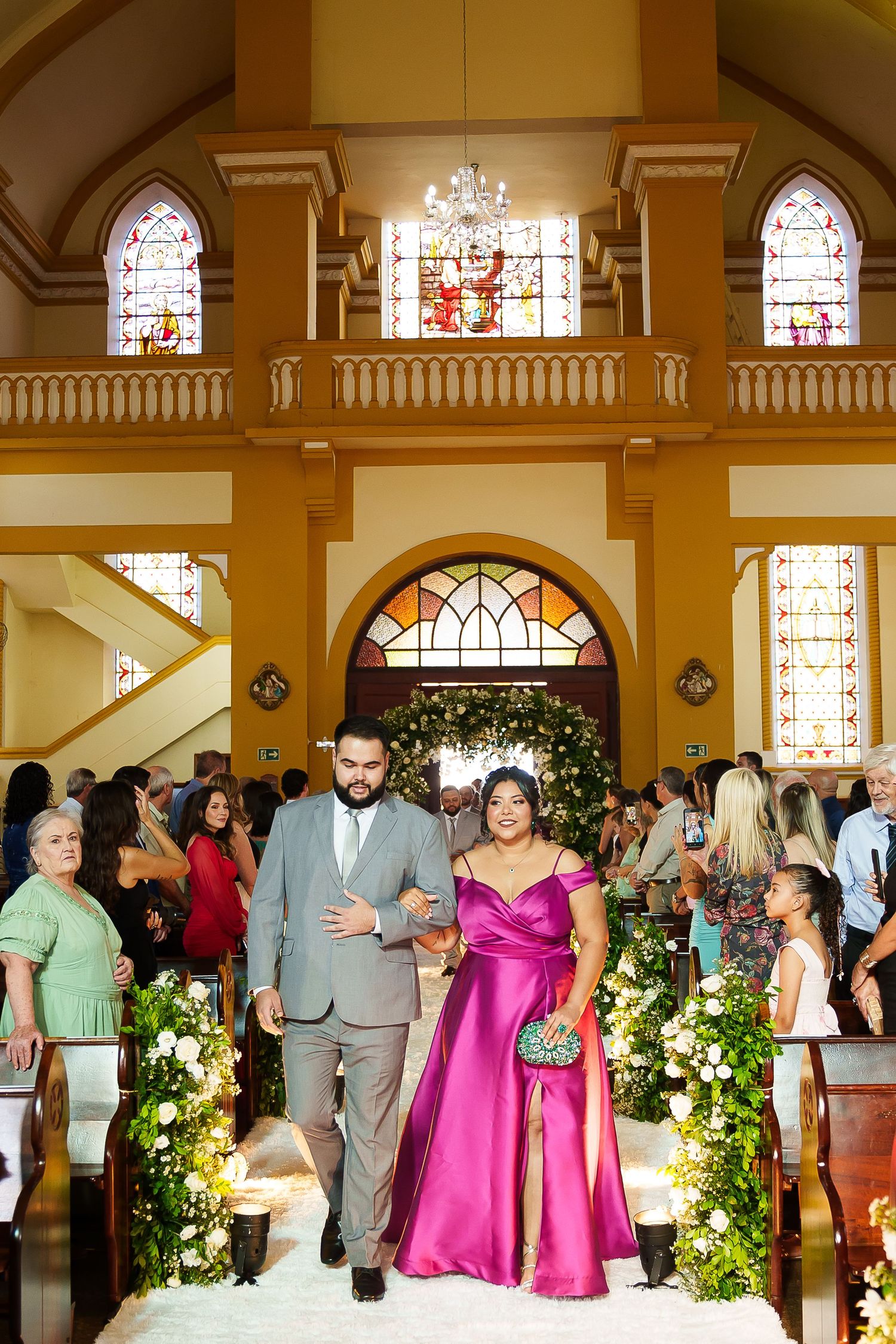 A couple walks down a church aisle, man in gray suit and woman in magenta gown, during a wedding ceremony.