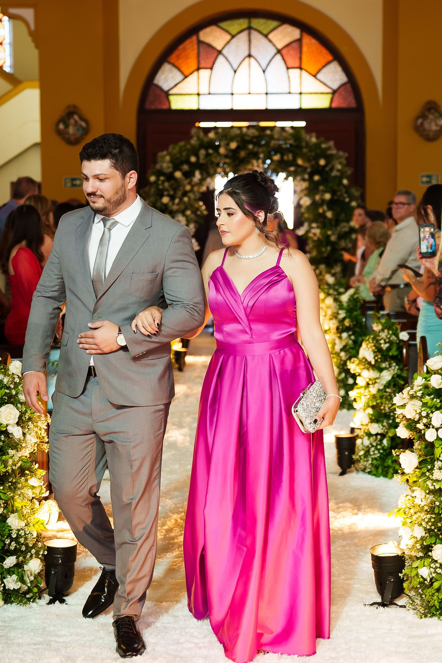 Couple walking down wedding aisle, man in gray suit, woman in pink satin gown, floral decorations.