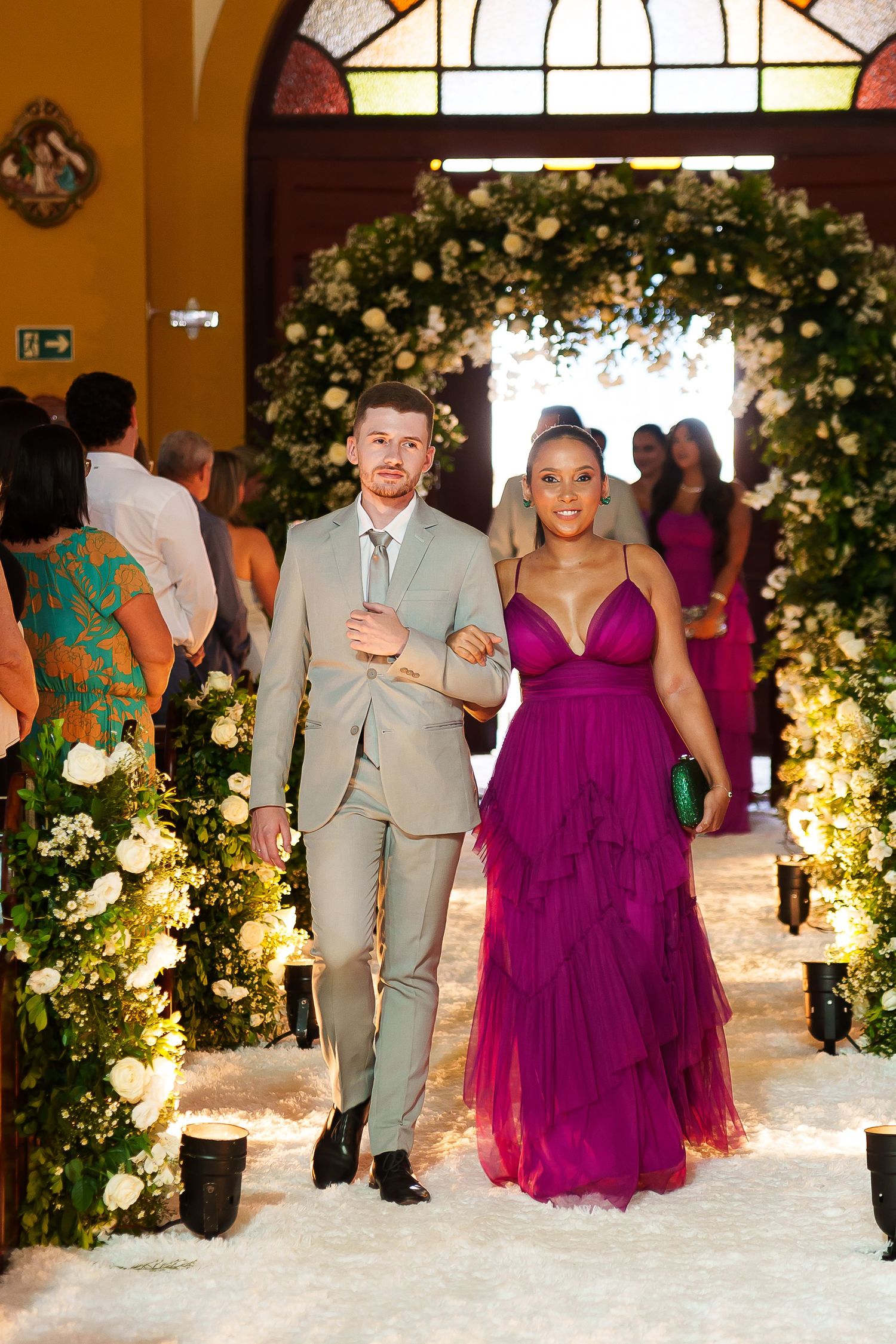 Couple walks down floral wedding aisle, man in gray suit, woman in purple tulle gown, elegant church ceremony.