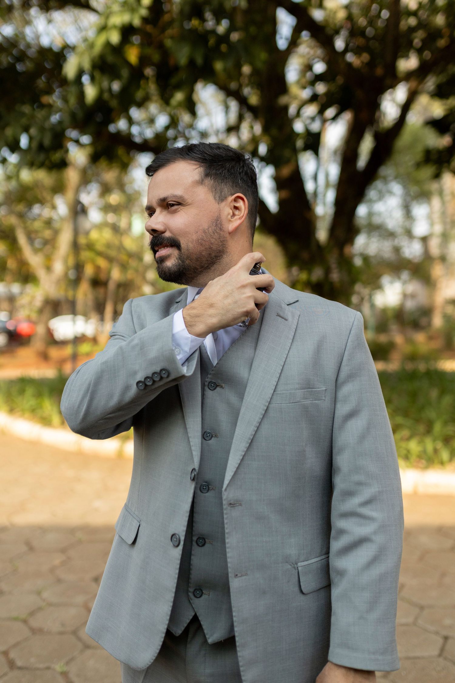 Bearded man in gray three-piece suit adjusting collar outdoors in a park setting.