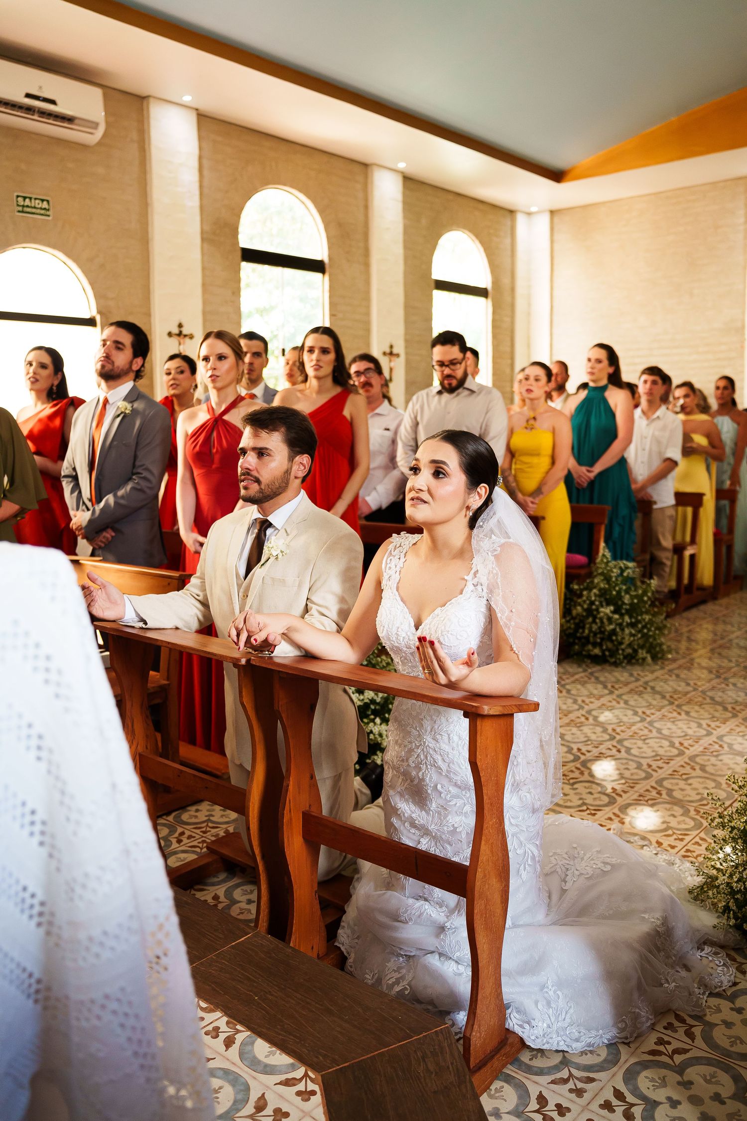 Bride and groom kneel at church altar during wedding ceremony with guests standing in pews behind them.