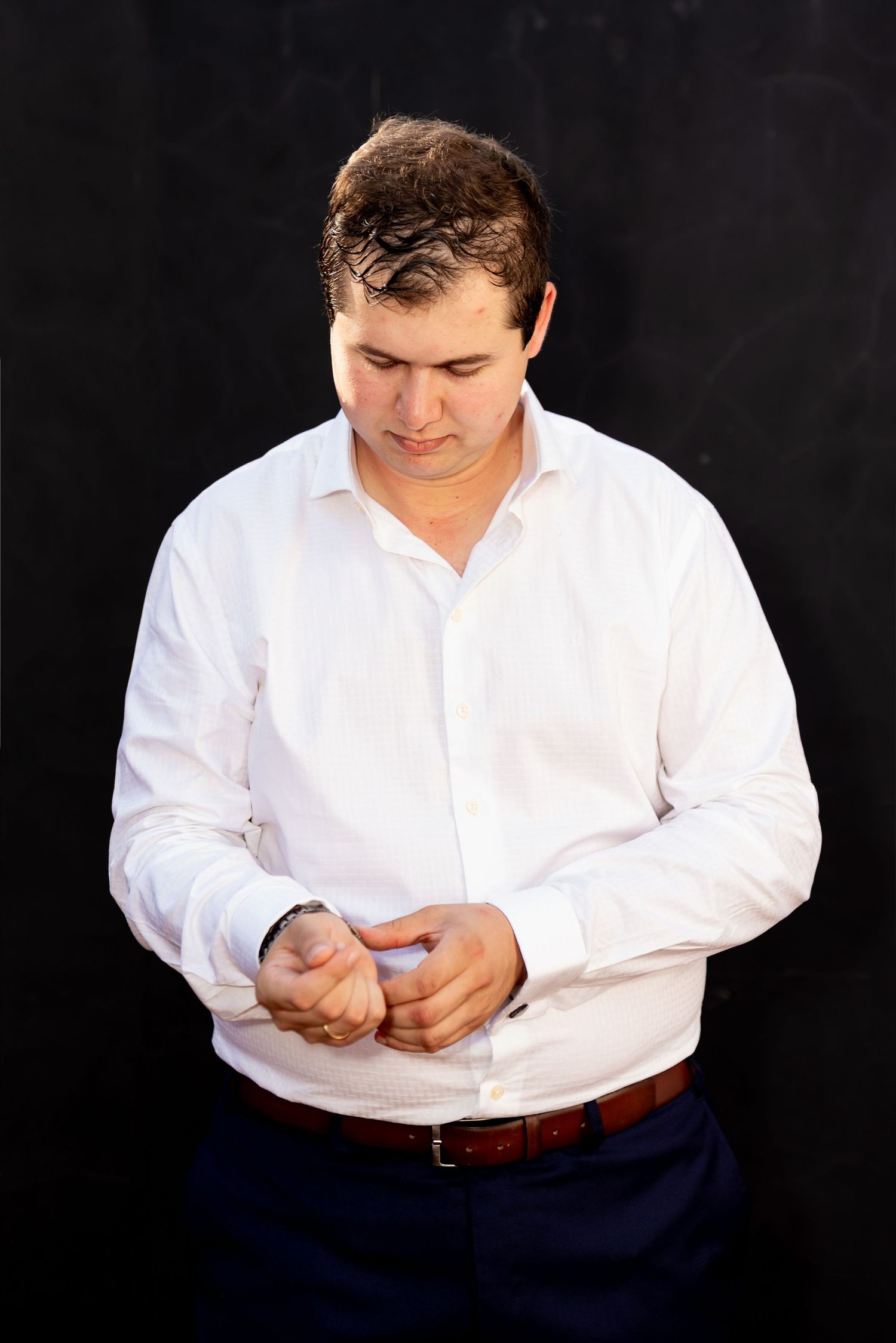 A young man in a white dress shirt adjusts his cufflinks against a dark background.