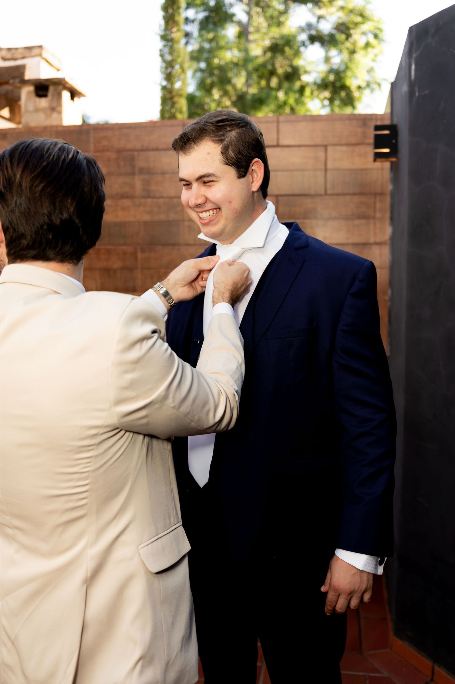 Groom in navy suit smiles as groomsman in tan suit adjusts his white tie outdoors before wedding ceremony.
