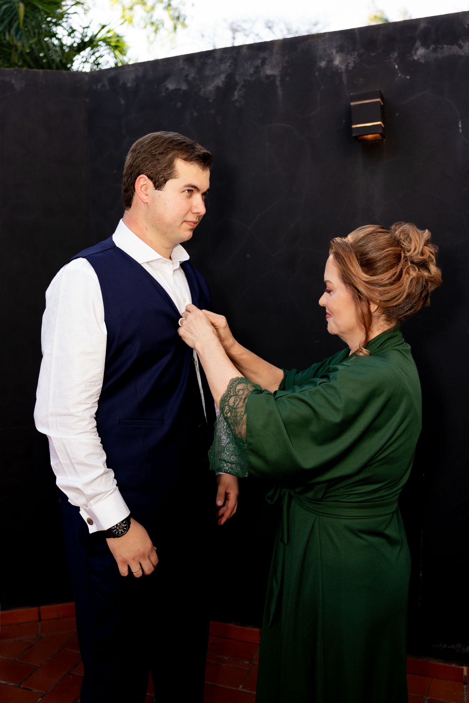 Woman in green dress adjusting boutonniere on man in navy vest and white shirt outdoors.