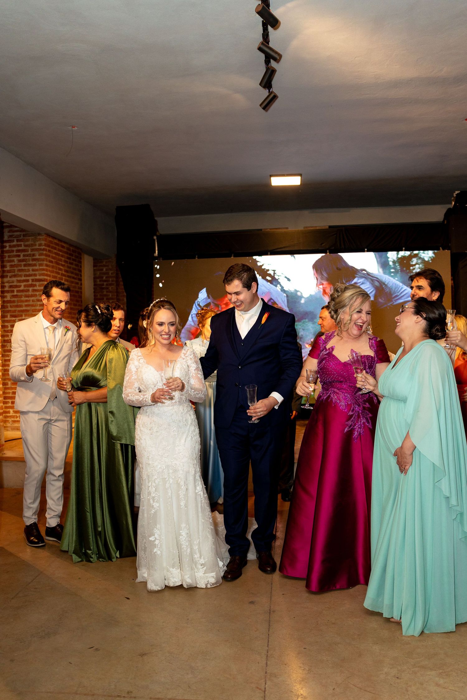 Wedding couple walks with family in colorful formal gowns at elegant indoor reception venue.